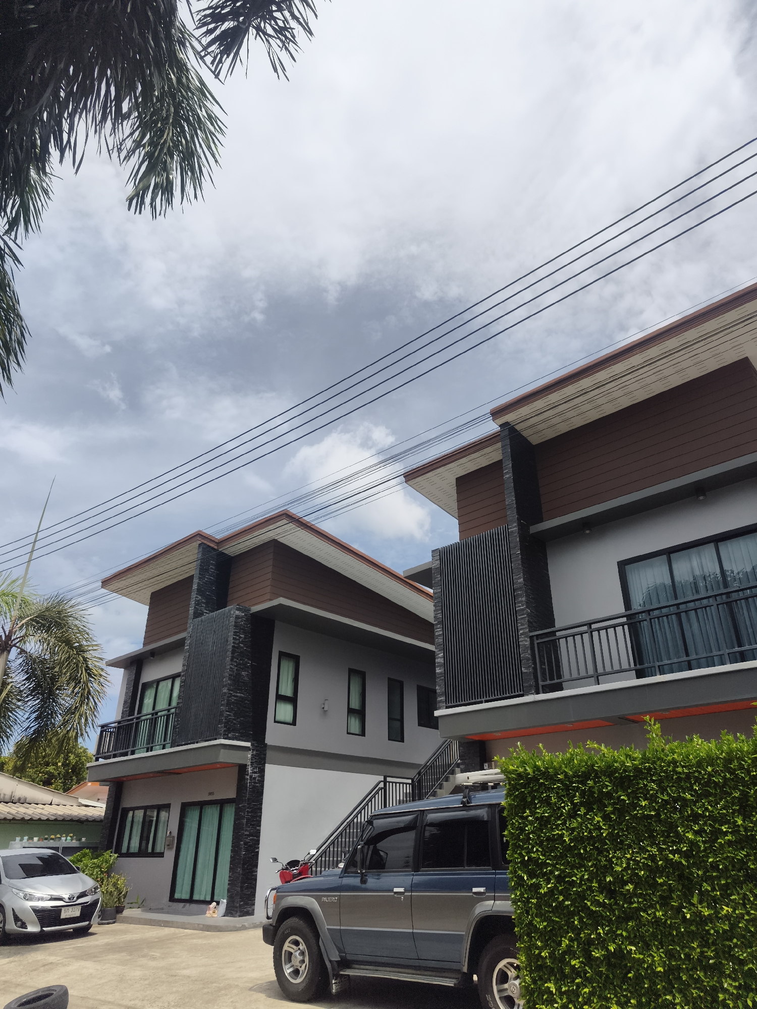 The image shows a modern, two-story house on a cloudy day in Ko Samui District, Thailand. The house features a gray facade with brown wooden accents on the upper portion and dark gray stone cladding around the entrance. Large windows and glass doors are visible on both floors. A black metal balcony with a railing is on the second floor.
In front of the house, a dark blue and silver SUV is parked on the concrete driveway. To the left of the SUV, a silver sedan is also parked. A red motorcycle is visible near the entrance to the house. A tall, well-maintained green hedge runs along the right side of the image. Palm trees are present in the foreground, with their fronds partially obscuring the sky, which is filled with white and gray clouds. Electrical wires run across the top of the frame. The scene appears to be a residential area.