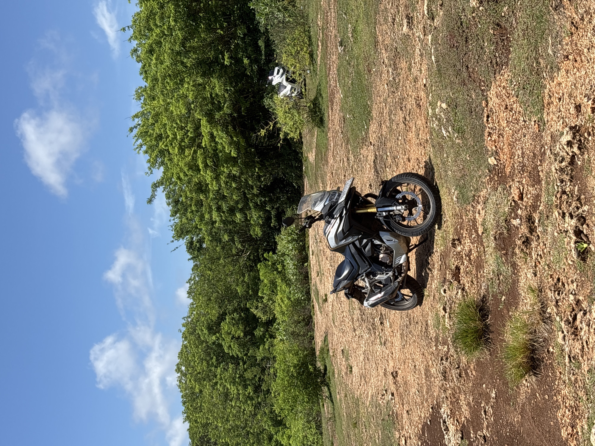 A black and silver motorcycle is parked on a brown, rocky, and grassy terrain. Dense green foliage borders the left edge of the frame, contrasting with a clear blue sky scattered with white clouds above. Further away, in the background, a small white object is visible among the trees. The scene is bathed in bright sunlight. This location may be near Uluwatu, Indonesia based on user information.