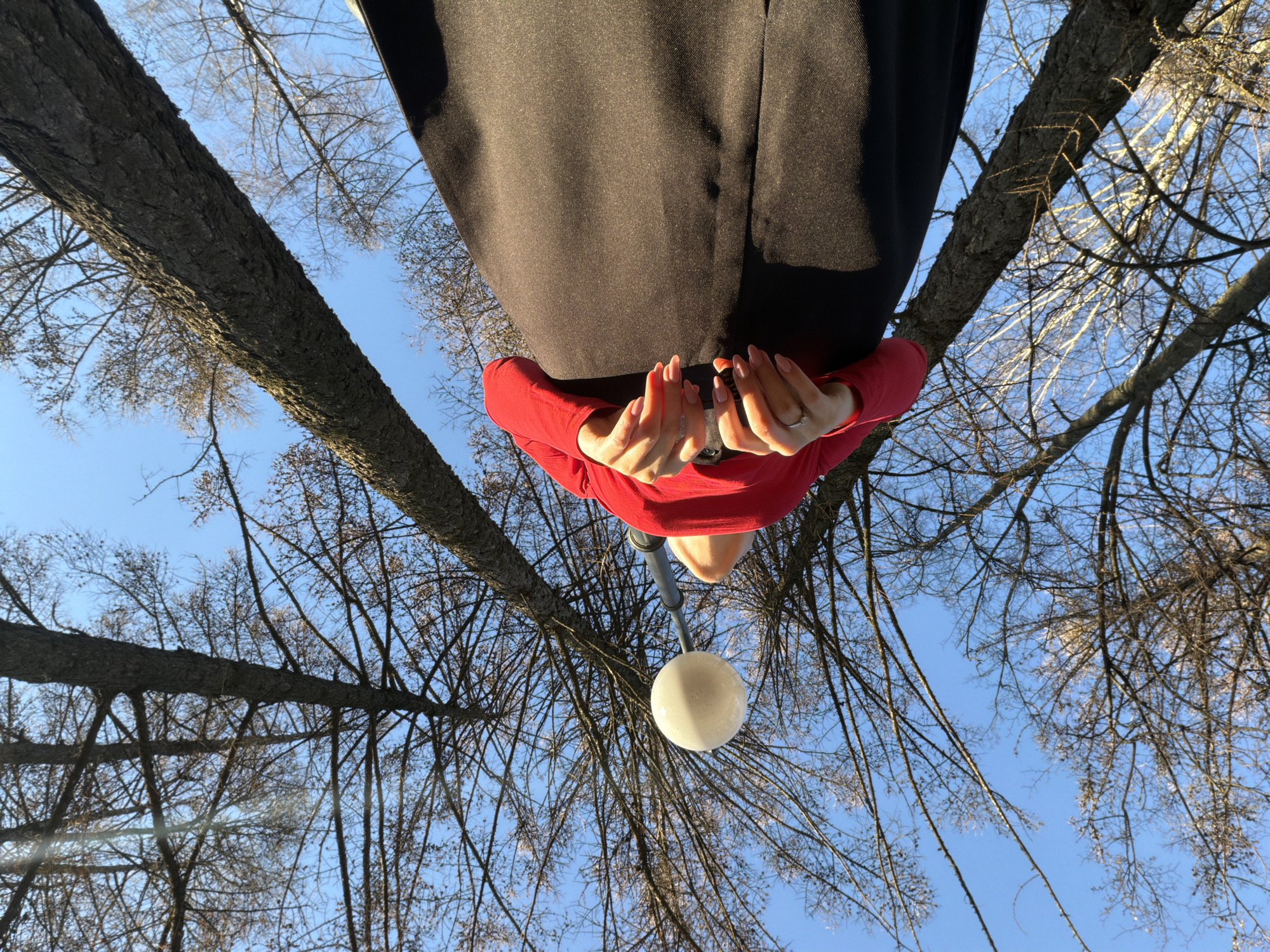 A low-angle shot captures a person holding a spherical object. The individual is wearing a red top and dark pants. The background consists of a blue sky and bare tree branches. The image was taken outdoors in what seems like the city of Izhevsk, Russia.