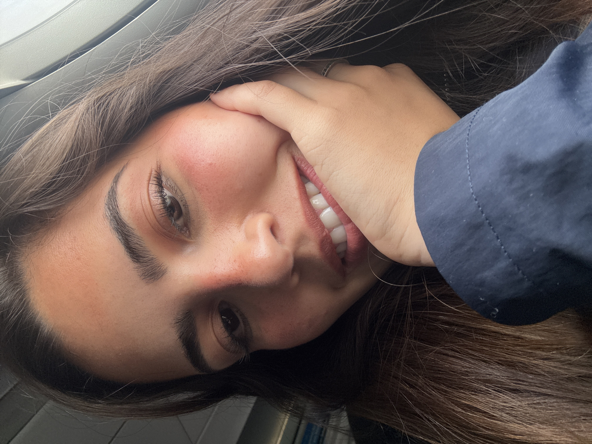 A young woman with brown hair, fair skin, and brown eyes is smiling in a close-up selfie. She is wearing a dark blue shirt and a ring is visible on her hand. The background appears to be the interior of a vehicle. The specific location within San Antonio, United States of America, is not determinable from the image.