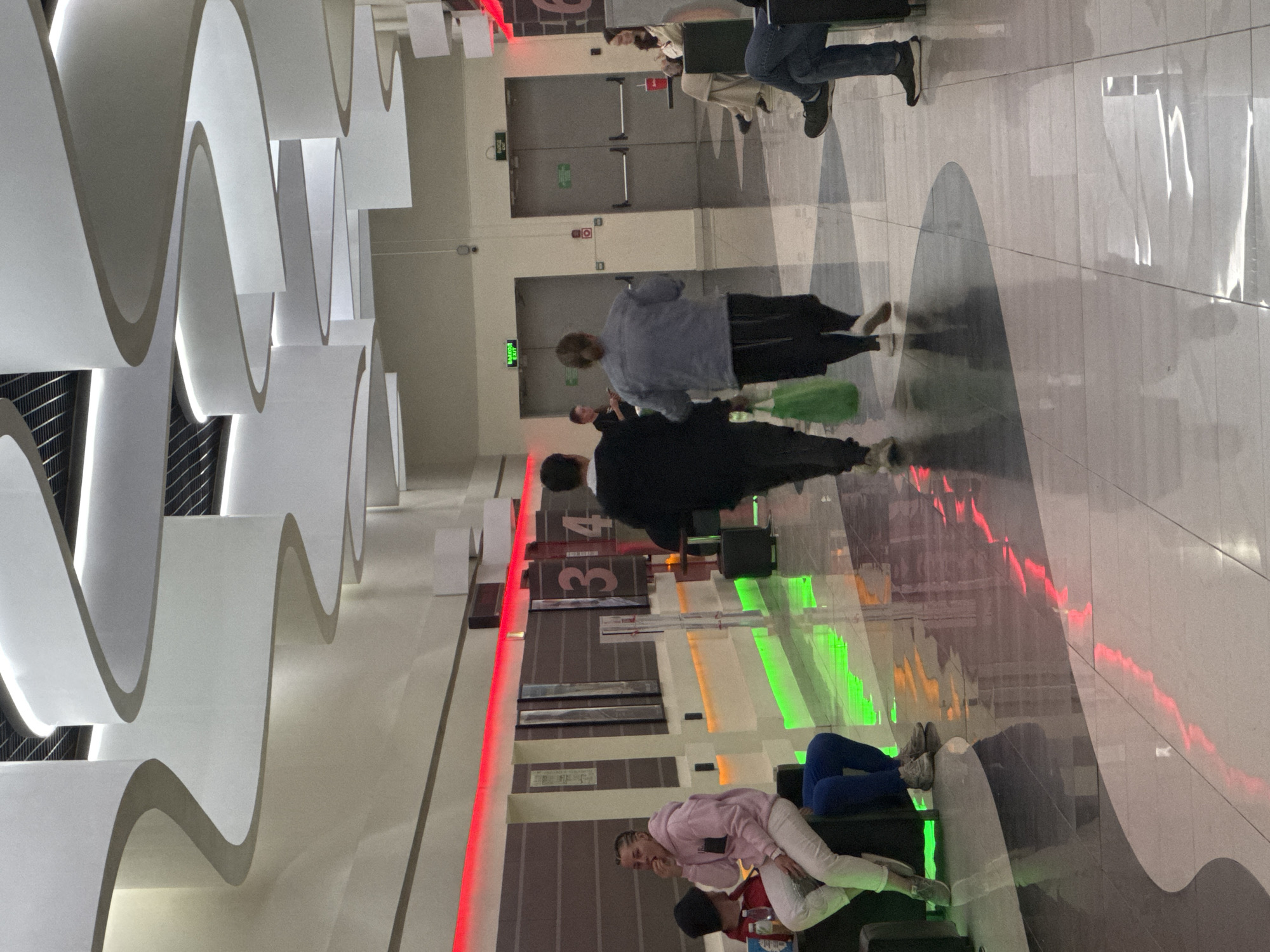 The image shows an indoor scene, possibly a transportation hub or a large public building, with several people present. The background features a distinctive architectural design with curved, white, wave-like structures along the ceiling. The floor is tiled with a polished surface reflecting light.
Several individuals are scattered throughout the scene. Some are standing and walking, while others are seated on what appears to be a set of steps or tiered seating area. Lighting is a mix of natural and artificial, with some colored lights, notably red and green, highlighting parts of the wall and floor. A sign with the number "34" is visible. There are also doors and exit signs in the distance. The location is in the city of Unknown, Unknown.