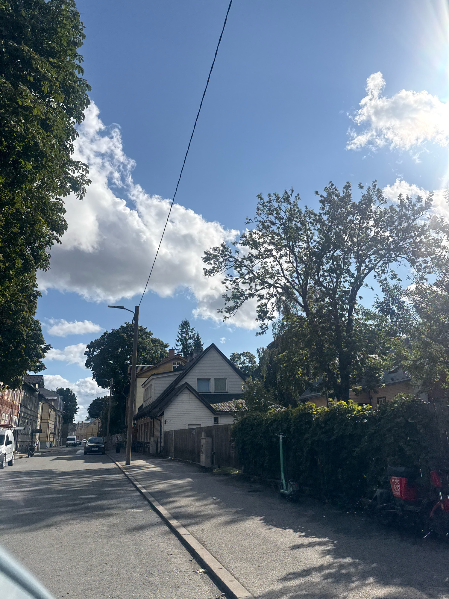 A street scene in Tallinn, Estonia, captures a sun-drenched day with dappled shadows on the asphalt road. Tall trees with dense green foliage line the street, interspersed with various residential buildings and a prominent street lamp. The sky is a vibrant blue, dotted with fluffy white clouds. A black car is parked on the side of the road, and further down, a white van is visible. In the foreground, on the right side of the street, a green electric scooter stands near a hedge, and a red motorcycle is parked behind it. The buildings in the background exhibit a mix of architectural styles, with some featuring wooden facades and others painted in lighter colors. The overall impression is one of a quiet urban neighborhood on a clear, sunny day.