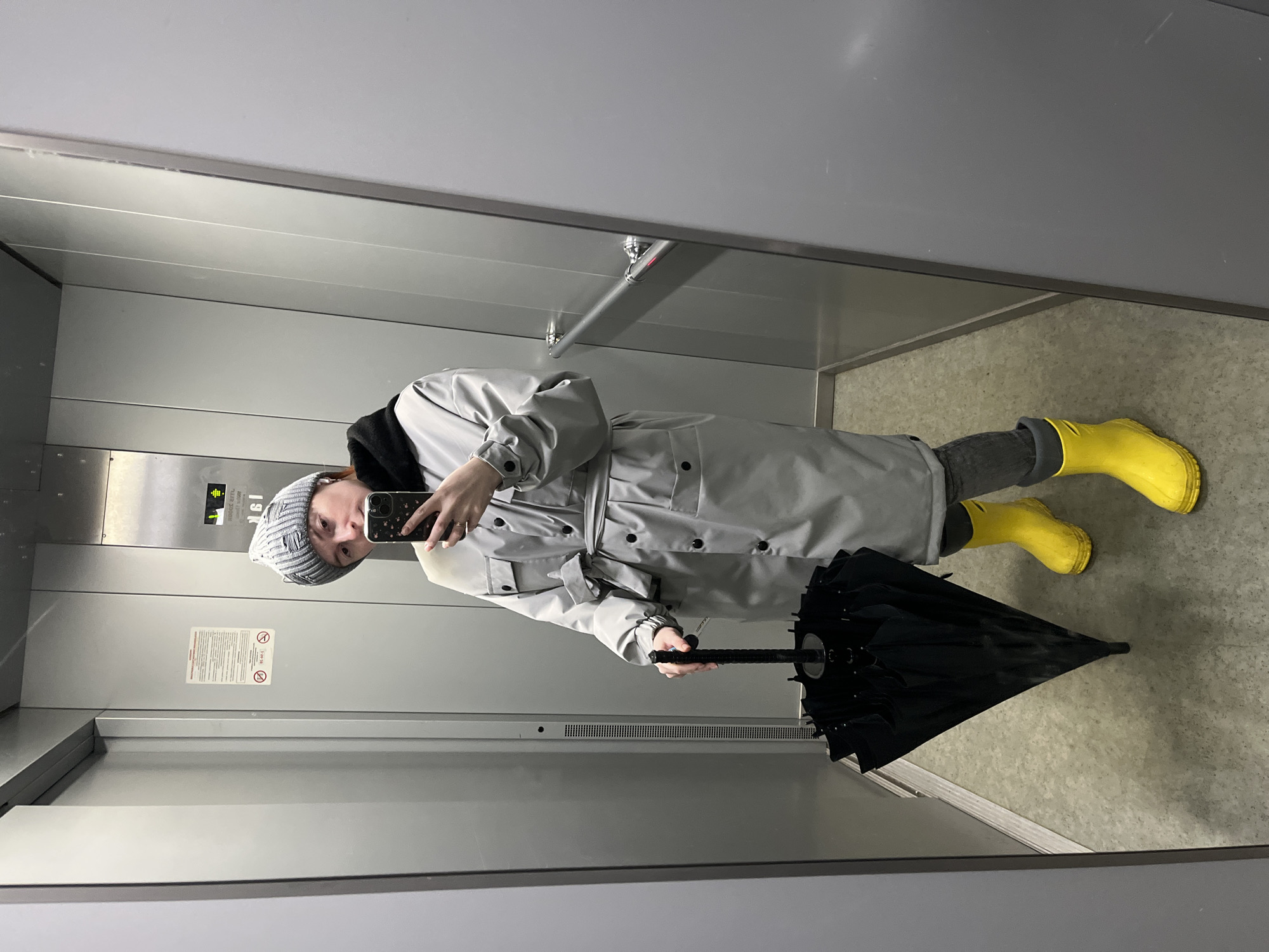 A person is reflected in a mirror in an elevator. The person is wearing a gray beanie, a black hoodie, a gray trench coat, gray pants, and yellow boots. They are holding a black umbrella in their left hand and a smartphone in their right hand. The elevator has gray walls and a gray floor. The location is in the city of Chaikovsky, Russia.