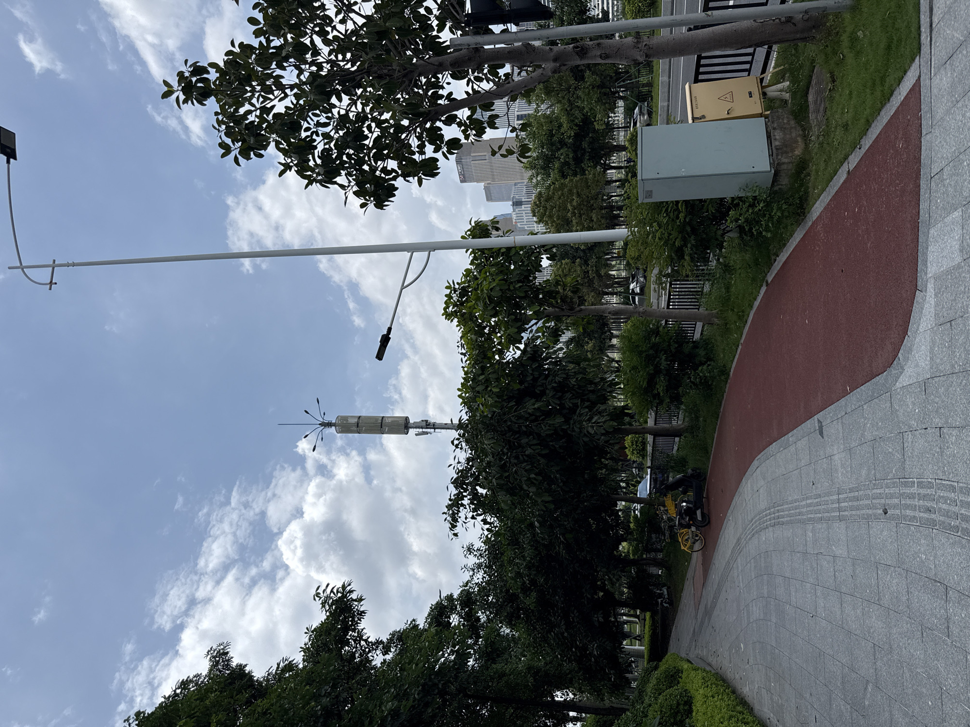 This image depicts a paved pathway in Haizhu District, China. The pathway is a combination of grey granite tiles and a red-colored surface, likely a bike lane or running track. To the left of the pathway are trees and manicured bushes. In the middle ground, a person is riding a yellow scooter. Further back, there are more trees, a cell tower, and a distant view of city buildings. A white lamppost with multiple lights is prominent on the right side of the frame, along with a light blue electrical box and a yellow hazard warning box. The sky is partly cloudy.