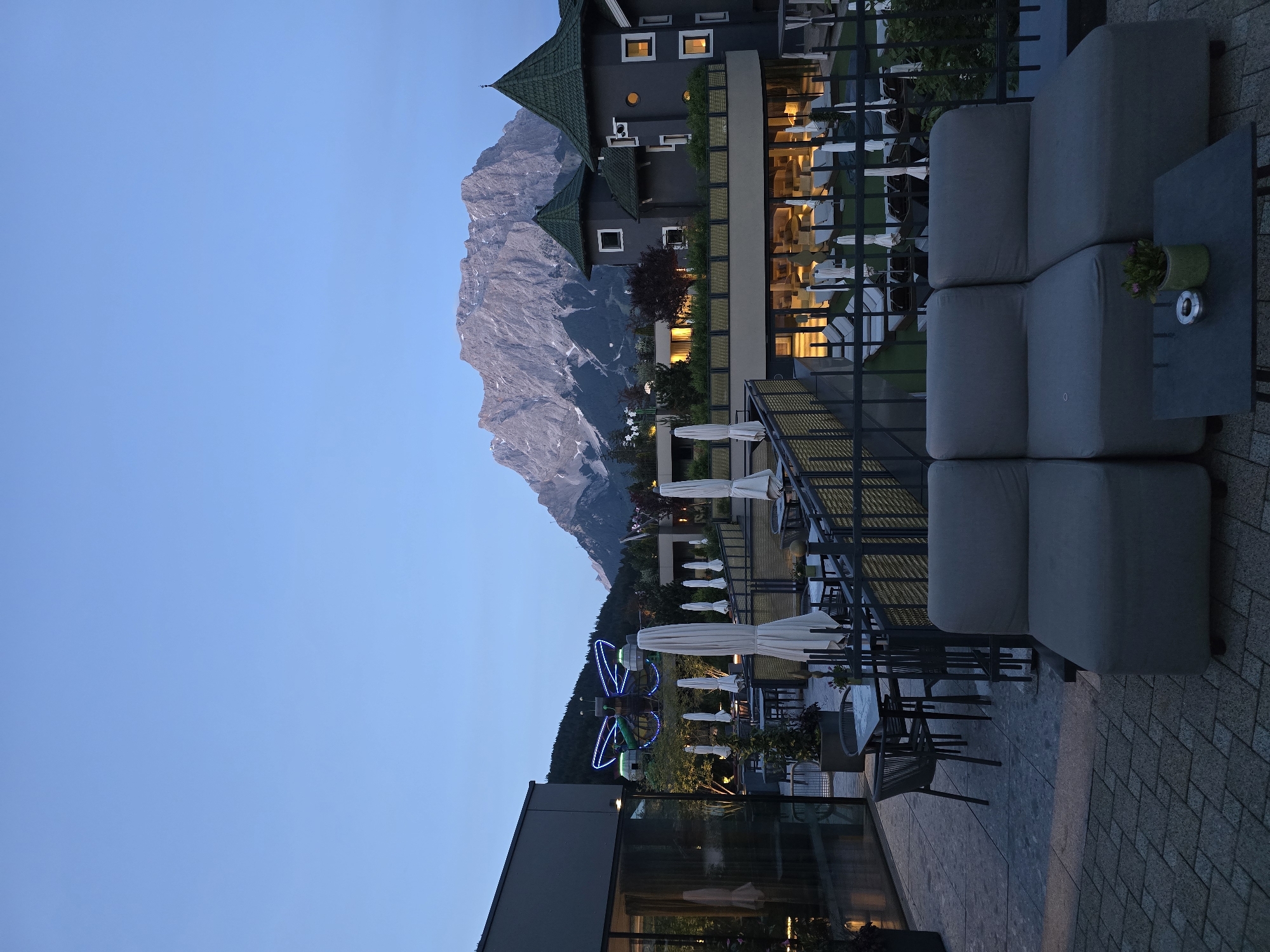 The image captures an evening view of an outdoor terrace or patio, likely belonging to a hotel or resort, set against a dramatic mountainous backdrop in Lermoos, Austria. The scene is illuminated by artificial lights against a twilight sky, indicating dusk or early night.
In the foreground, a comfortable outdoor lounge area is visible with several modular grey sofas and small grey tables. One table holds a green potted plant and a silver ashtray. Black metal railings separate the patio from a lower section. To the left, a modern building with large glass windows is partially seen.
The midground features a striking architectural structure, presumably the main hotel building, characterized by dark grey walls and distinctive green, multi-tiered pagoda-style roofs. Its windows glow with warm yellow light. Further down the gentle slope from the hotel, a large, brightly lit decorative structure, resembling a butterfly or insect, glows in blue and green hues. Several closed white outdoor umbrellas are scattered across the lower patio areas.
Dominating the background is a massive, rugged mountain, appearing grey and imposing with visible rocky textures and some lighter patches, possibly snow or bare rock. The lower slopes of the mountain are covered in dark green trees. The sky above is a clear, deep blue, devoid of clouds.
No people are visible, lending a serene and tranquil atmosphere to the luxurious outdoor setting. There is no discernible activity taking place, but the arranged seating suggests a space designed for relaxation or social gatherings. No text is visible in the image.