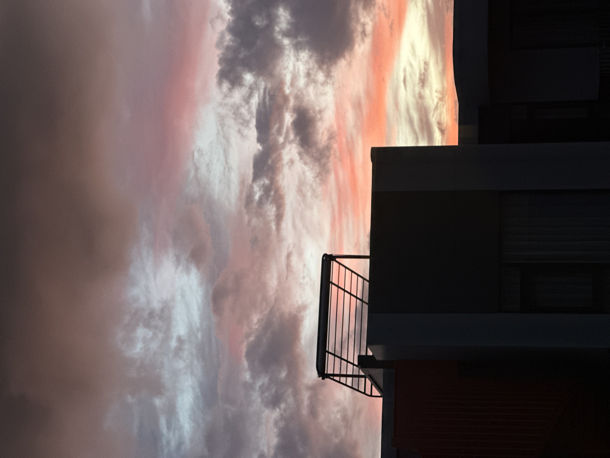 A silhouetted rooftop structure, possibly a satellite dish or solar panel mount, is visible against a dramatic sunset sky. The sky is filled with wispy clouds in hues of pink, orange, and purple, with streaks of lighter blue and gray. In the foreground, the dark silhouettes of building rooftops create a stark contrast to the vibrant sky. The scene appears to be an urban or suburban environment, possibly in Choeng Thale, Thailand, given the context. No people are visible in the image.