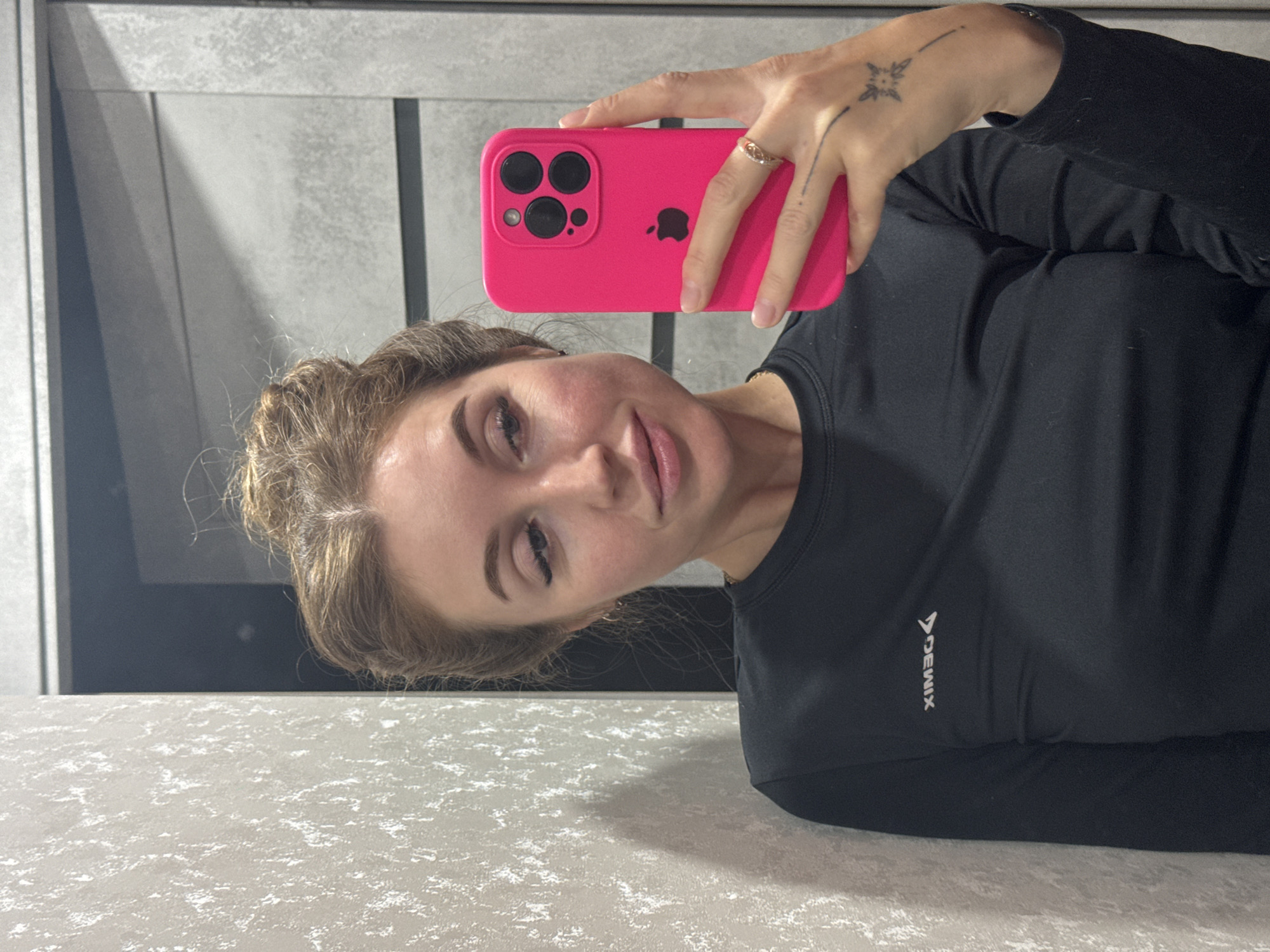 A fair-skinned woman is taking a selfie with a pink iPhone. She is wearing a black long-sleeved shirt with a logo that reads "DEMIX". The environment appears to be an indoor space with a gray patterned surface in the foreground. Behind her is a wall with gray paneling. The image was taken in the city of Chaikovsky, Russia.