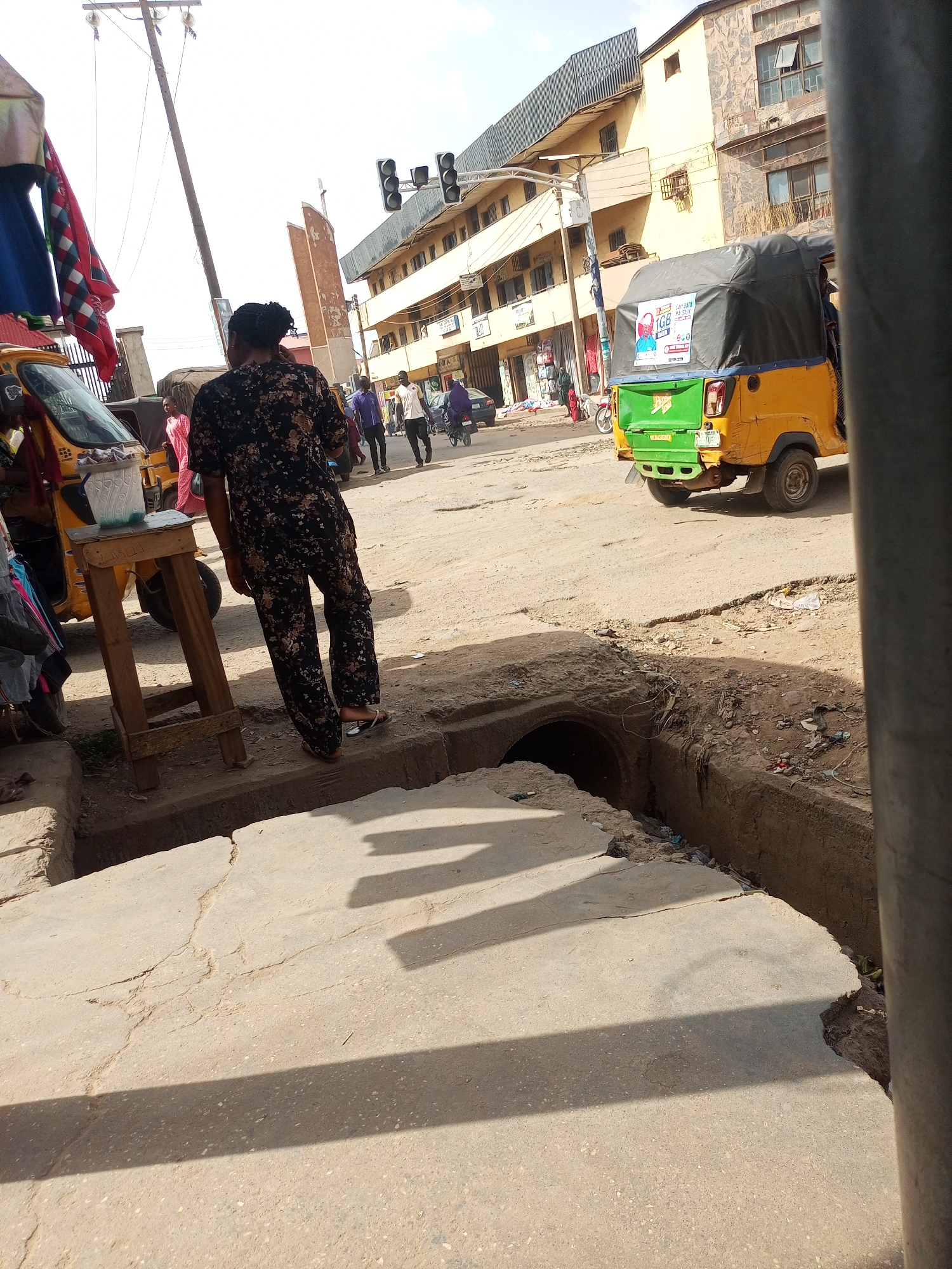 The image depicts a street scene in Maiburiji, Nigeria. A woman in a floral print jumpsuit stands near an open drainage ditch along the edge of a concrete sidewalk. A yellow and green auto-rickshaw (tricycle) is partially visible on the right side of the frame. Another auto-rickshaw is on the left, near a makeshift wooden table with items on it. Pedestrians are walking on the street in the background. Buildings line the street, and a traffic light is visible above the street. The road appears dusty and slightly uneven. Shadows are cast on the sidewalk, and the drainage ditch contains debris.