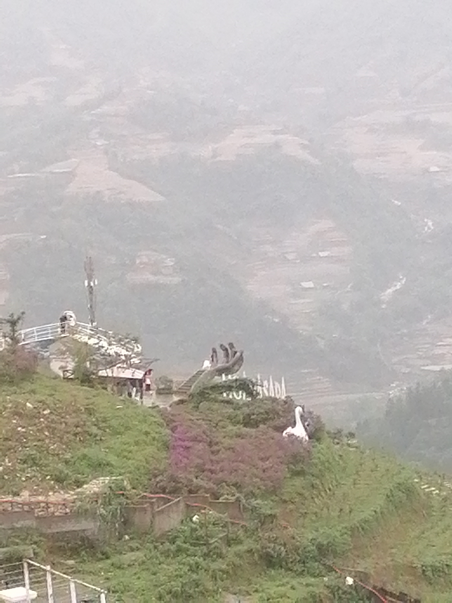 The image shows a hillside scene in Sa Pa Ward, Vietnam. The foreground features a green, sloping hillside with vegetation, including some pink flowering plants and a white swan statue. There's a concrete structure or retaining wall visible, as well as a partial view of a white fence in the lower left. A platform area with multiple people is visible; it has a lookout point or viewing area and what appears to be a cell tower. The background reveals a hazy mountain landscape, likely part of the Hoàng Liên Son mountain range near Sa Pa Ward. The visibility is low due to what could be fog or atmospheric conditions.