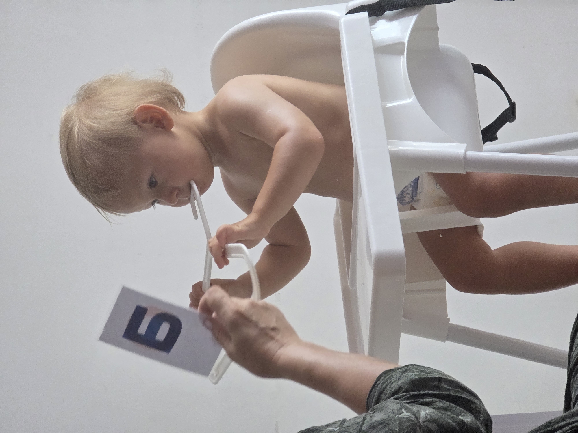 A toddler, likely a boy due to his blonde hair and bare chest, is seated in a white high chair. He is wearing a diaper. The toddler is holding a white plastic object to his mouth and looking to the left. To the left of the toddler, a person's hands are visible, holding a white card with a dark blue letter "Б" on it. The background is a plain white wall, suggesting an indoor setting. The image is cropped and does not provide any information about the city of Sochi, Russia.