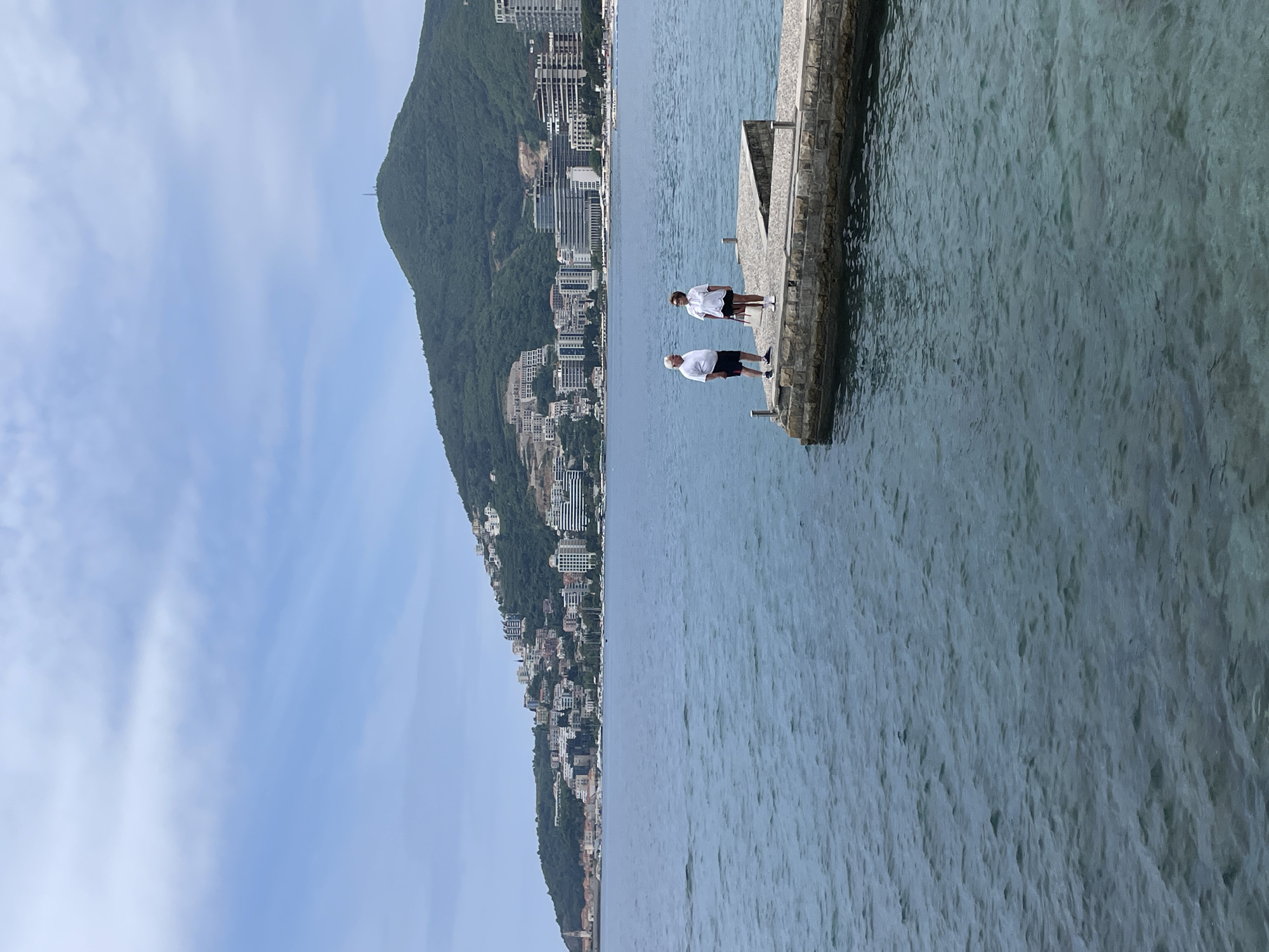 Here is a detailed description of the image:
Two people stand on a stone pier in the city of Budva, Montenegro. One person is an older male in a white shirt, black shorts, and a white face mask. The other individual is a younger person in a white shirt, black shorts, and a white face mask. The pier extends into the clear blue water. Behind the pier and to the left of the frame, a mountain rises covered with lush green vegetation. Buildings, likely residences or hotels, are densely packed along the slope of the mountain. The sky above the mountain is blue with some white clouds. The scene suggests a coastal urban environment.