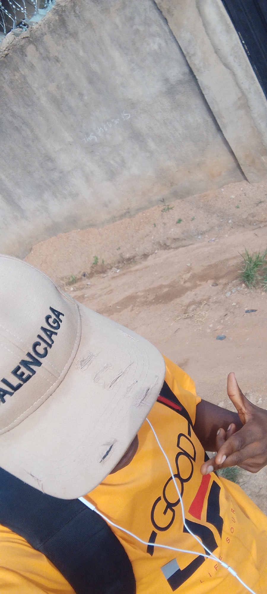 In Madalla, Nigeria, a person wearing a tan baseball cap with "BALENCIAGA" embroidered on it and a yellow t-shirt with the word "GOOD" is visible. The cap has rips on the front. The person is also wearing black socks and a black backpack. White earbuds are connected to the t-shirt, suggesting the person is listening to music. The background consists of a grey concrete wall with some chalk graffiti that reads "this hou=15", and a dirt path with sparse vegetation. A hand is visible gesturing with a thumb and index finger.