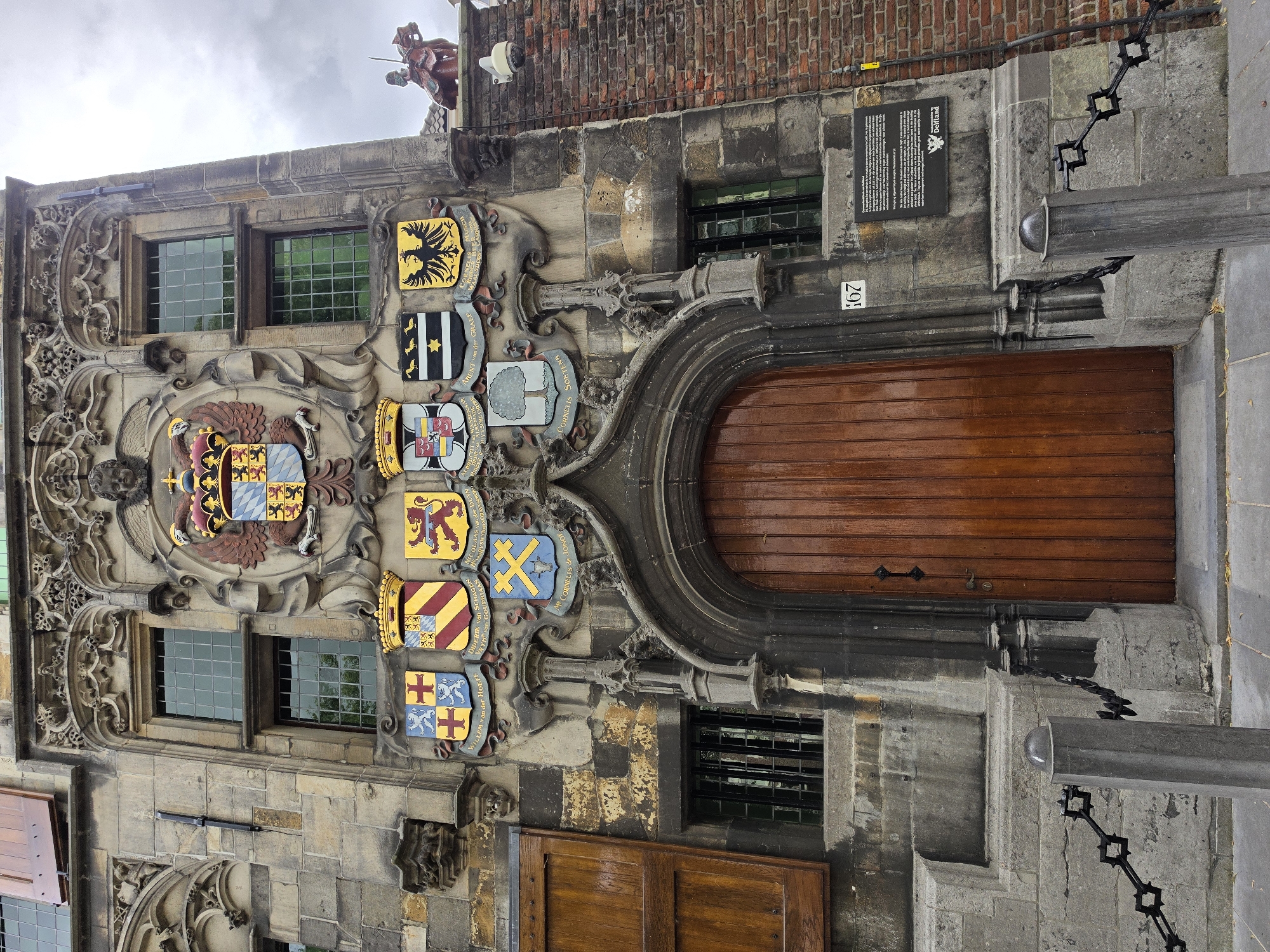The image shows the facade of a building with a large wooden door and several coats of arms above it. The building appears to be made of stone, with decorative carvings and architectural details. There are also some windows with green panes visible. To the right of the door, there is a brick wall and a statue of a person on horseback. A sign with the number "167" is posted next to the door. The sky is overcast. The building is located in Delft, Netherlands, as indicated by the Delfland logo on the sign. The sign itself is a historical marker, providing information about the building's history and its connection to the Delfland water board.