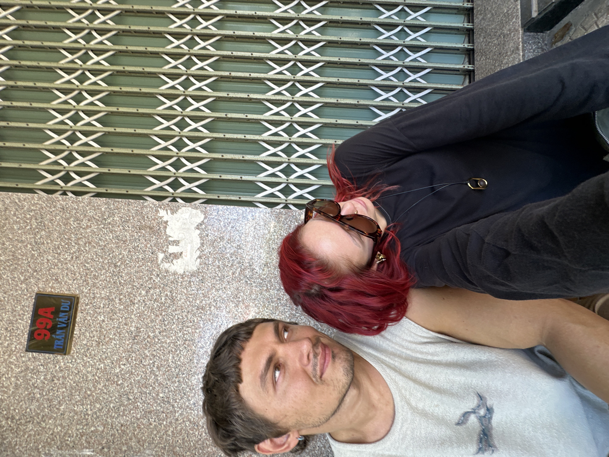 A man and a woman, possibly a couple, are sitting next to each other outside. The man, with short brown hair and a light-colored t-shirt, is looking to his left. The woman, with bright red hair and sunglasses, is leaning her head on the man's shoulder, looking down. She is wearing a black long-sleeved shirt with a distinctive pendant necklace. Behind them, a building wall with a sign that reads "99A TRAN VAN DU" is visible, along with a green collapsible gate. The overall setting appears to be an urban environment, possibly a residential area or a storefront in Đà Nẵng, Vietnam, given the Vietnamese address.