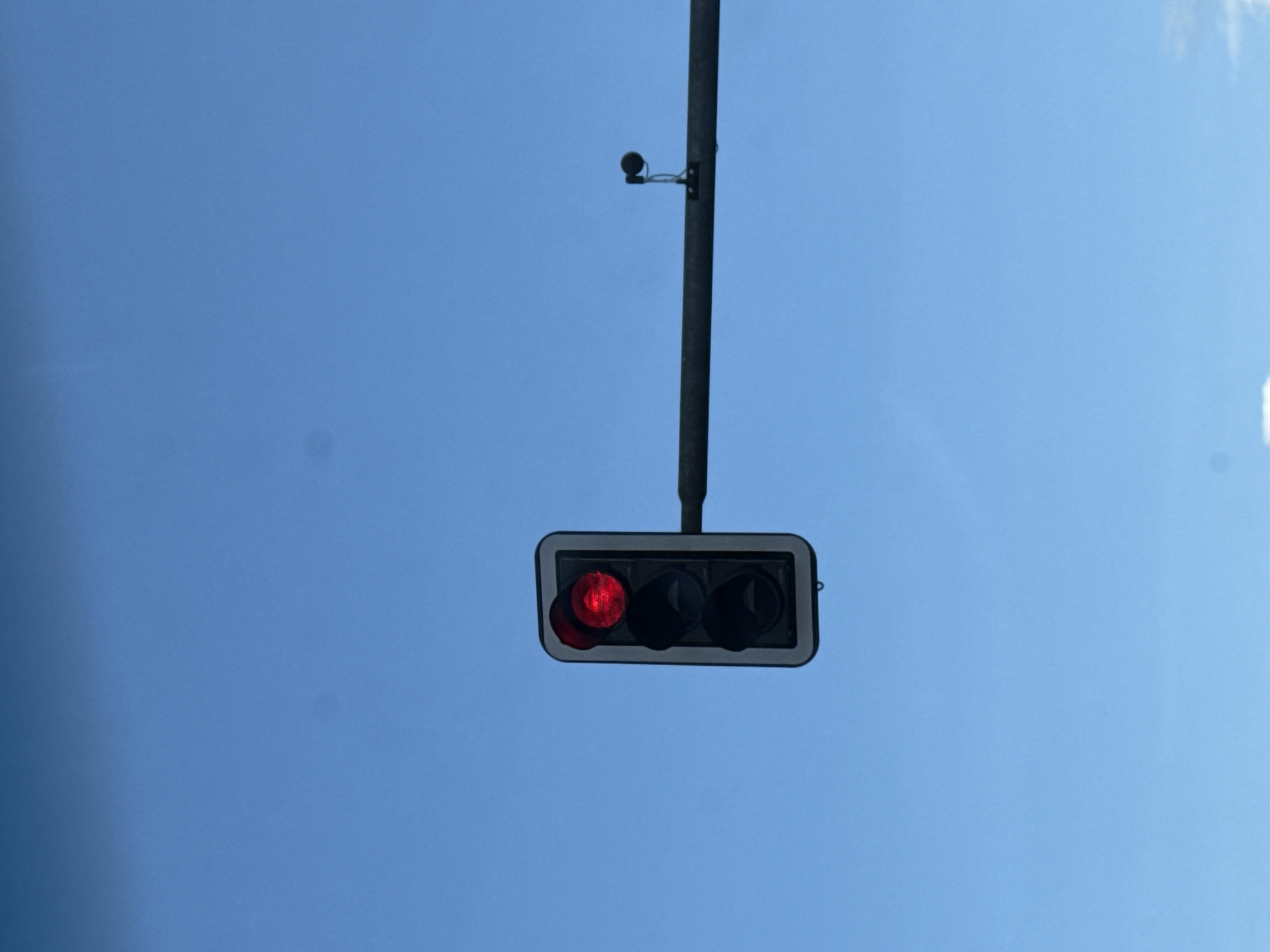 A vertical traffic signal with a lit red light is positioned to the left of a horizontal pole that extends across the frame. A small, black, circular device is attached to the pole to the right of the traffic signal. The background is a clear blue sky. The image is undated. The location of this scene is Herzberg, Germany.