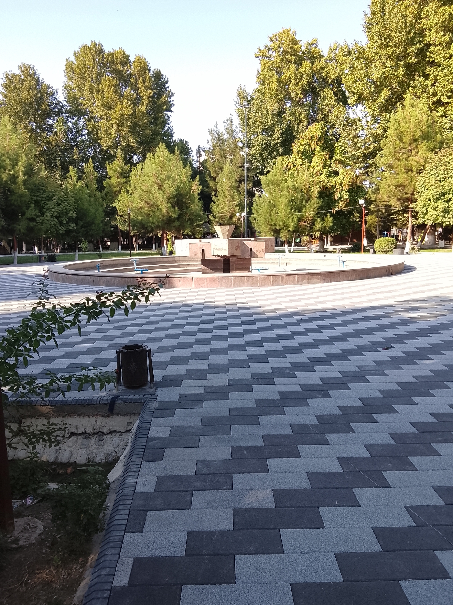 A paved public square features a large, circular, dry fountain at its center. The fountain has a raised, multi-tiered stone structure in the middle. The pavement is a geometric pattern of dark gray and lighter gray rectangular tiles. Lush green trees with dense canopies surround the square, with a clear sky overhead. In the foreground, to the left, a young tree with sparse leaves grows, and a dark trash receptacle stands beside the pavement. The scene appears to be a well-maintained park or plaza.