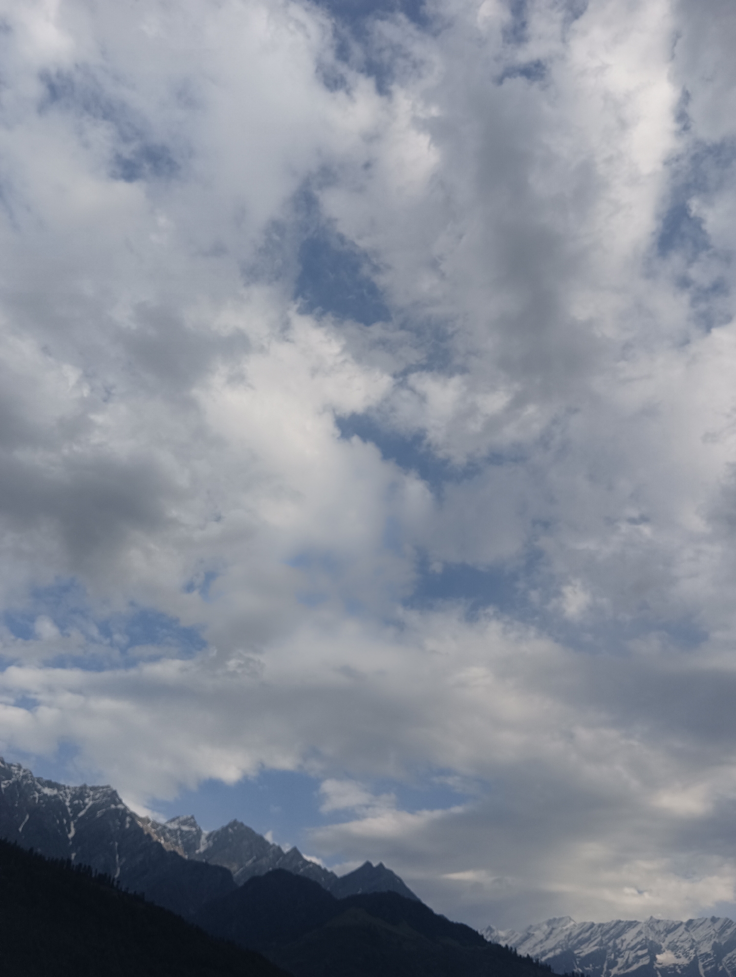 The image depicts a mountainous landscape with a dramatic sky filled with clouds. In the foreground, dark, forested slopes descend from the left side of the frame. The middle ground is dominated by jagged mountain peaks, some of which are capped with snow, reflecting the light. The background is a vast sky, a mosaic of white and grey clouds interspersed with patches of blue sky. The lighting suggests it is either early morning or late afternoon. The scene is characteristic of the mountainous regions surrounding Vashisht, India, known for its natural beauty and trekking opportunities.
