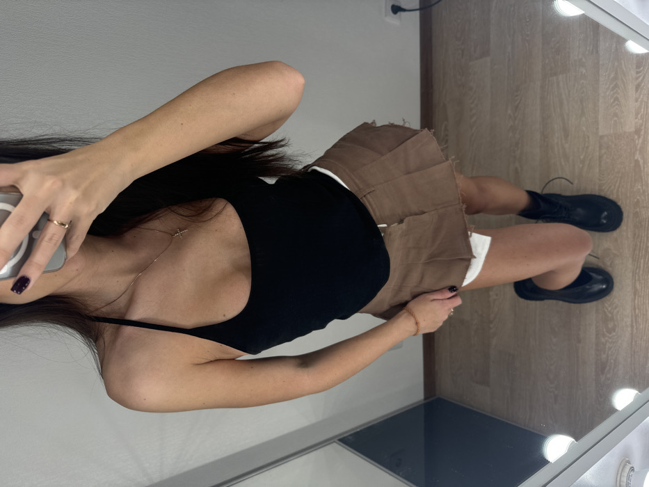 The image shows a full-body mirror selfie of a person, likely a young woman, indoors. The person has long, dark hair that falls over their shoulders and chest. They are wearing a black spaghetti-strap tank top or camisole. Around their neck, a delicate gold chain with a small cross pendant is visible. On their left hand, a simple gold ring is worn on the ring finger, and their nails are painted a dark color, possibly deep red or purple. The person is holding a light-colored smartphone to take the picture, partially obscuring their face.
Below the black top, they are wearing a brown, pleated mini-skirt that appears to have raw or unfinished edges. White fabric, possibly undershorts or a lining, peeks out from beneath the skirt. On their right wrist, a thin, light-colored bracelet is visible. The person is wearing black lace-up combat boots.
The setting appears to be an indoor room with a light brown wood laminate floor. The wall behind the person is a plain light grey or off-white, with a black electrical outlet visible on it. The selfie is taken in a large mirror, the frame of which is visible on the right side of the image, featuring multiple exposed round light bulbs, characteristic of a vanity mirror. The lighting in the room is bright, likely from these mirror lights. The overall pose suggests a casual outfit check or fashion shot.