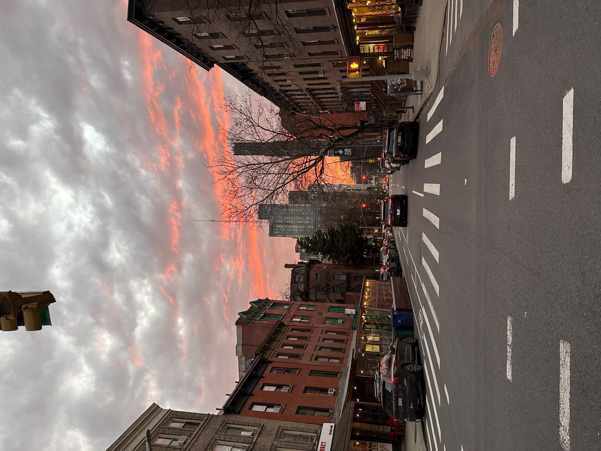 The image displays a street scene on Court Street in Carroll Gardens, Brooklyn, New York City, United States, viewed northwards at dusk. The sky presents a dramatic sunset, characterized by widespread gray clouds interspersed with intense red and orange light dominating the horizon and upper mid-ground.
The foreground features a two-way asphalt street with white-painted crosswalk stripes and dashed lane markings. On the left side, a dark gray Volvo SUV (NY license plate ELH 7136) is parked, adjacent to an outdoor dining enclosure with string lights, accompanied by blue and green refuse bins. A continuous row of three-to-five-story brick and brownstone buildings lines the left side of the street, housing ground-level commercial establishments, one displaying a partial "MARKET" sign. One building further down features distinctive green trim.
On the right, similar multi-story buildings extend down the block. The closest building on this side houses a warmly lit commercial establishment, "VERO BUONO LIQUOR & WINES SPIRITS," with decorative string lights. A single individual is partially visible on the sidewalk near a brown newsstand. Bare deciduous trees with distinct branch patterns are scattered along both sides, while a dark evergreen tree stands on the right.
Vehicles are both parked and in motion on the street. Several dark-colored cars, including a prominent dark sedan, are traveling away from the viewer. Overhead, a traffic light displays a green signal. A pedestrian traffic light on the right shows a red "Don't Walk" hand symbol. In the distance, a cluster of modern skyscrapers, indicative of Downtown Brooklyn, rises against the bright red and orange sky, with some illuminated windows and an antenna visible above. A red manhole cover is visible in the street on the right. The scene depicts an active urban environment transitioning from day to night.