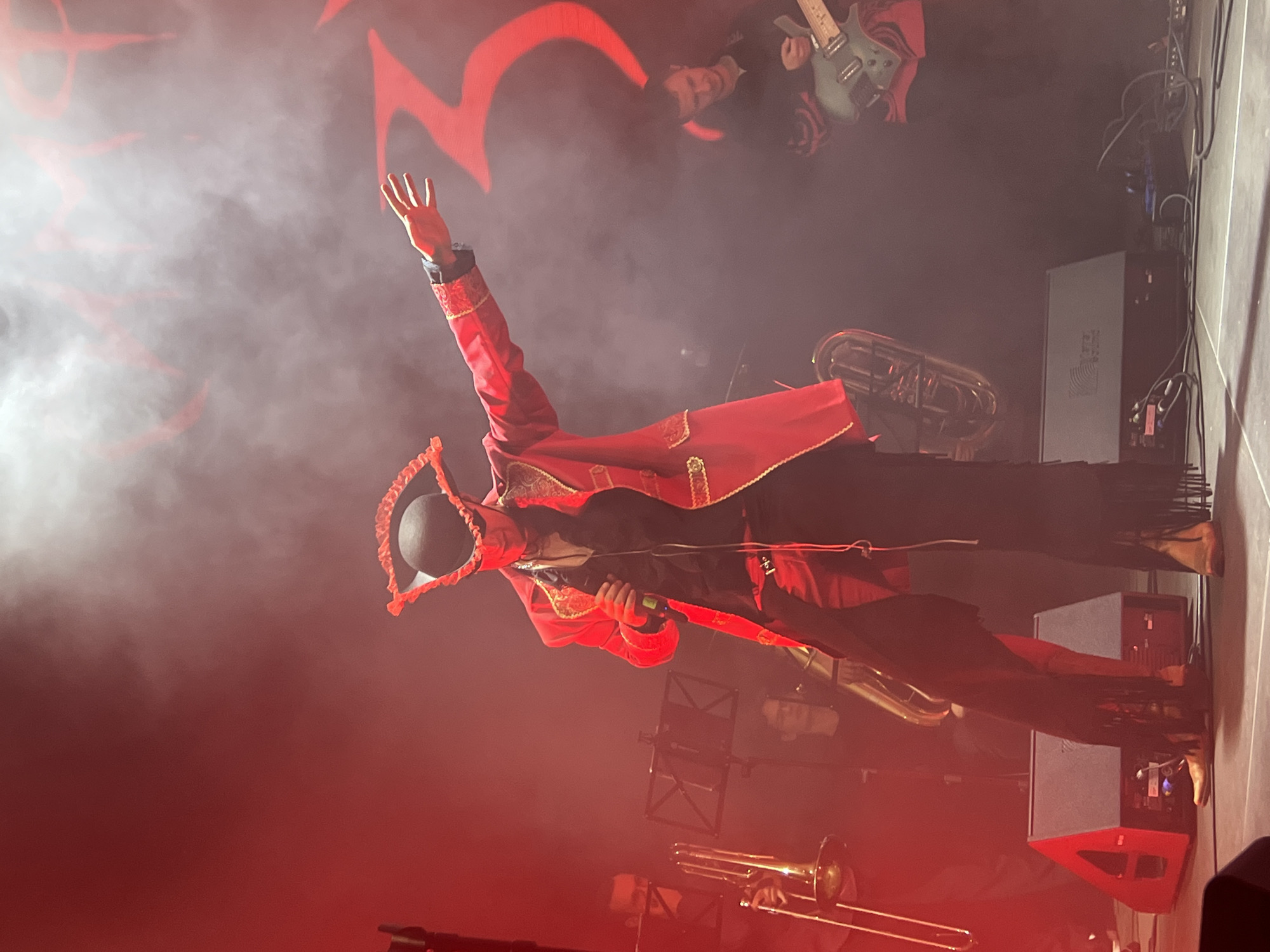 The image shows a performer on stage in a red jacket, black pants with fringe, and a hat, with one arm raised. The performance appears to be lit with red stage lights. There are musical instruments including a trombone and a tuba visible on stage. There is a guitarist partially visible in the upper right of the image. A speaker cabinet is located stage right. The image was taken at a performance in Saint Petersburg, Russia.