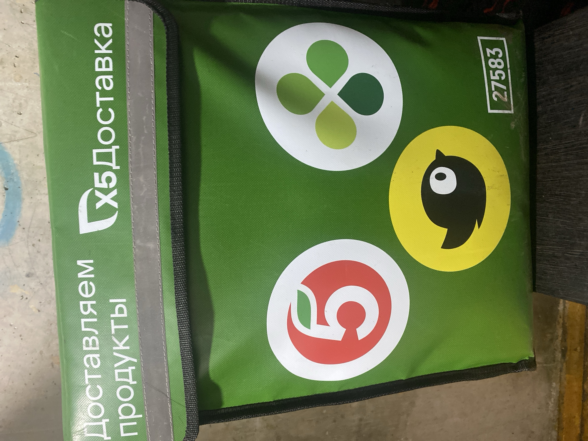 A green delivery bag with "Доставляем продукты" and "Х5 Доставка" printed on it is shown. The bag features three circular logos: a red circle with a white "5" and a green leaf, a white circle with a clover-like design in shades of green, and a yellow circle with a stylized black bird. A white rectangle containing the number "27583" is visible in the lower right corner. The bag appears to be placed on a wooden surface, possibly a shelf or table, against a textured, grey background that resembles a concrete wall. The overall scene suggests a delivery service operating in an urban environment.