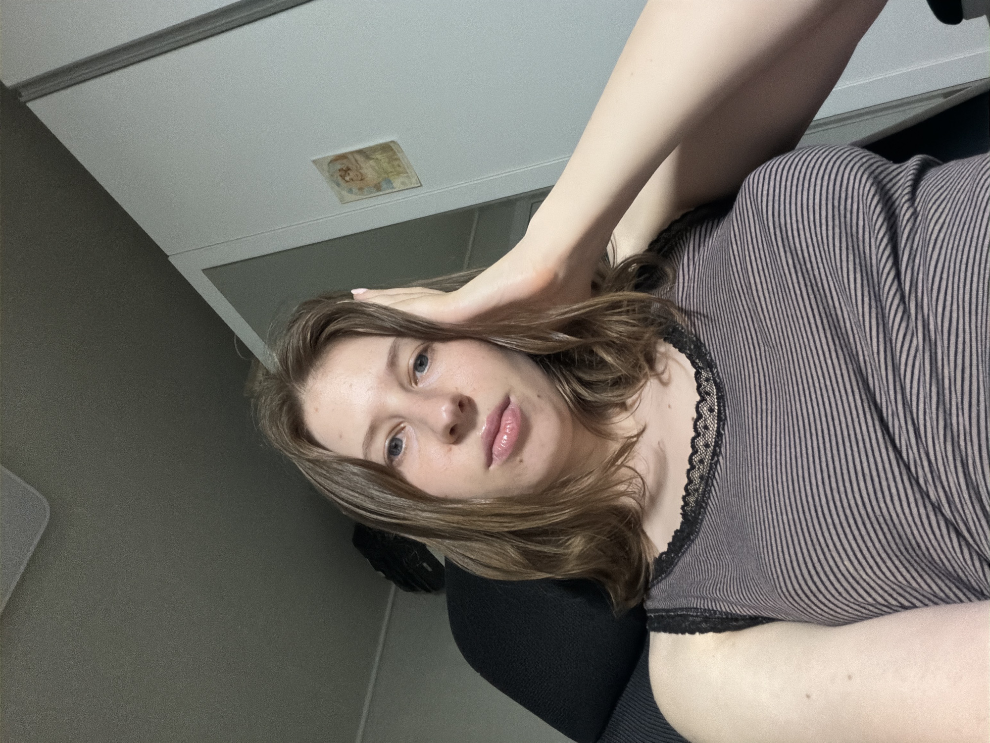 A young woman with shoulder-length brown hair is seated in a black office chair. She is wearing a horizontally striped tank top with black lace trim. Her right hand is resting on her head. She appears to be indoors, with a light-colored wall and a closet door visible in the background. There is also a framed picture on the wall. The scene is illuminated by artificial light. The woman's expression is neutral, and she is looking directly at the camera. The image appears to have been taken in Dongducheon-si, South Korea.