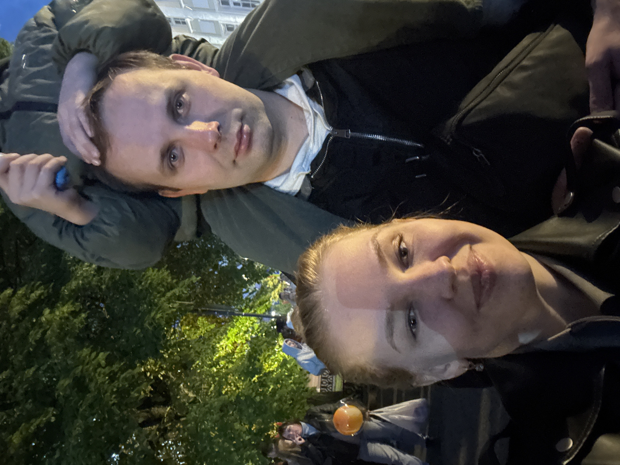 The image depicts two individuals in an outdoor setting. A man lies on his back, cradling his head with one arm, while a woman's face is positioned close to the camera. The background includes foliage and illuminated areas suggesting a street or park environment. No information is available regarding the location in the city of Podosenikha, Russia. Other individuals are visible in the background.