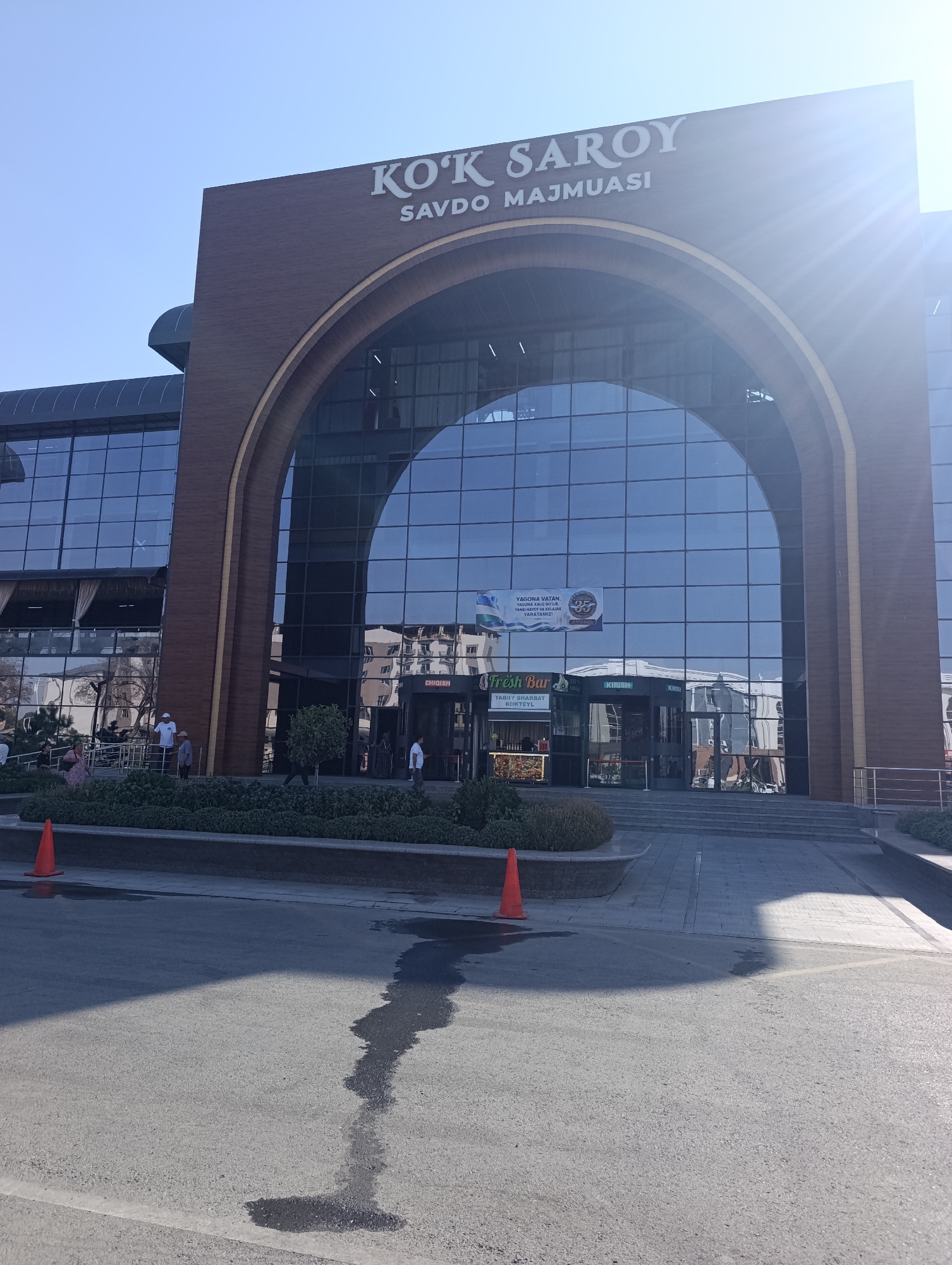 The image shows the exterior of the Kok Saroy shopping center in Jizzakh, Uzbekistan. The building features a large arched glass entrance, with a wooden facade above it bearing the text "KOK SAROY SAVDO MAJMULIS". Several people are visible in front of the building, some walking and others standing. There is also a "Fresh Bar" sign to the left of the entrance. The ground in front of the building is paved, with a dark wet patch on the asphalt. An orange traffic cone is visible on the left side of the image. The sky is clear and blue.
