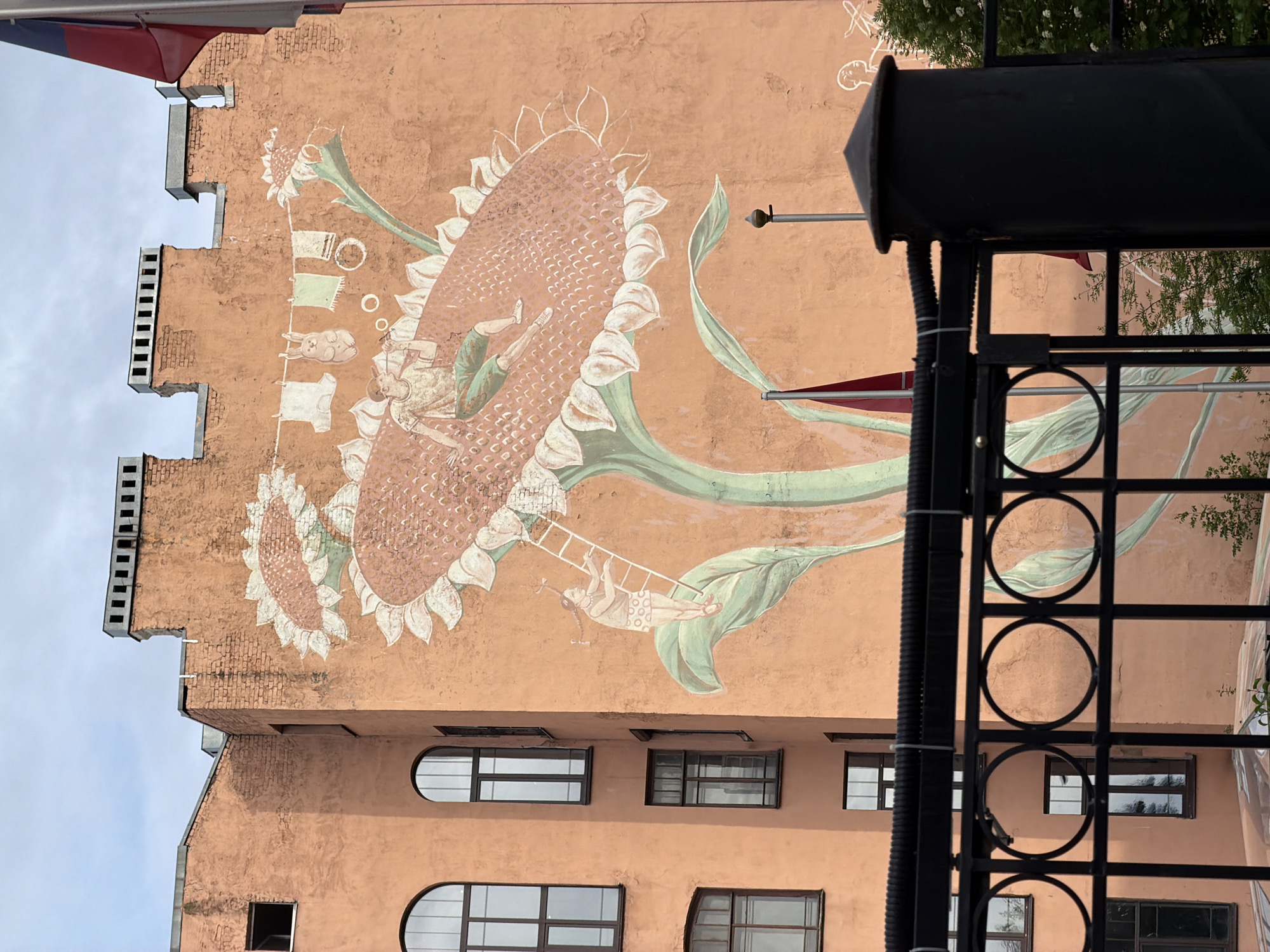 A large, vertically oriented photograph captures a whimsical mural on the side of a peach-colored building in Saint Petersburg, Russia, under an overcast sky.
The central feature is a massive mural depicting oversized sunflowers. Two prominent sunflowers dominate the frame; one large, detailed with small squares on its head, and a smaller one to its left. Several figures interact with these fantastical plants: one individual is depicted relaxing or sunbathing on the head of the largest sunflower, with their legs casually extended. Another smaller figure, possibly a child, climbs a white ladder extending from a sunflower leaf towards the main bloom. To the left, a clothesline hangs between sunflower elements, displaying items like shirts and a small rabbit figure. Faint chalk-like outlines of figures are also visible further up.
The building itself has a distinctive architectural style, characterized by a stepped, crenellated roofline visible on the left side. Below the mural, multiple windows of varying shapes are visible, including arched and rectangular designs with dark frames.
In the right foreground, a black metal structure, possibly a balcony railing or gate, partially obstructs the view. This structure features decorative circular cutouts and a dark conical cap. Beyond it, green foliage from trees can be seen. A red and blue banner or flag is partially visible in the top left corner.
The scene conveys a dreamlike, imaginative atmosphere against an urban backdrop, captured during daytime with soft, diffused light due to the cloudy conditions. No visible text is present in the image.