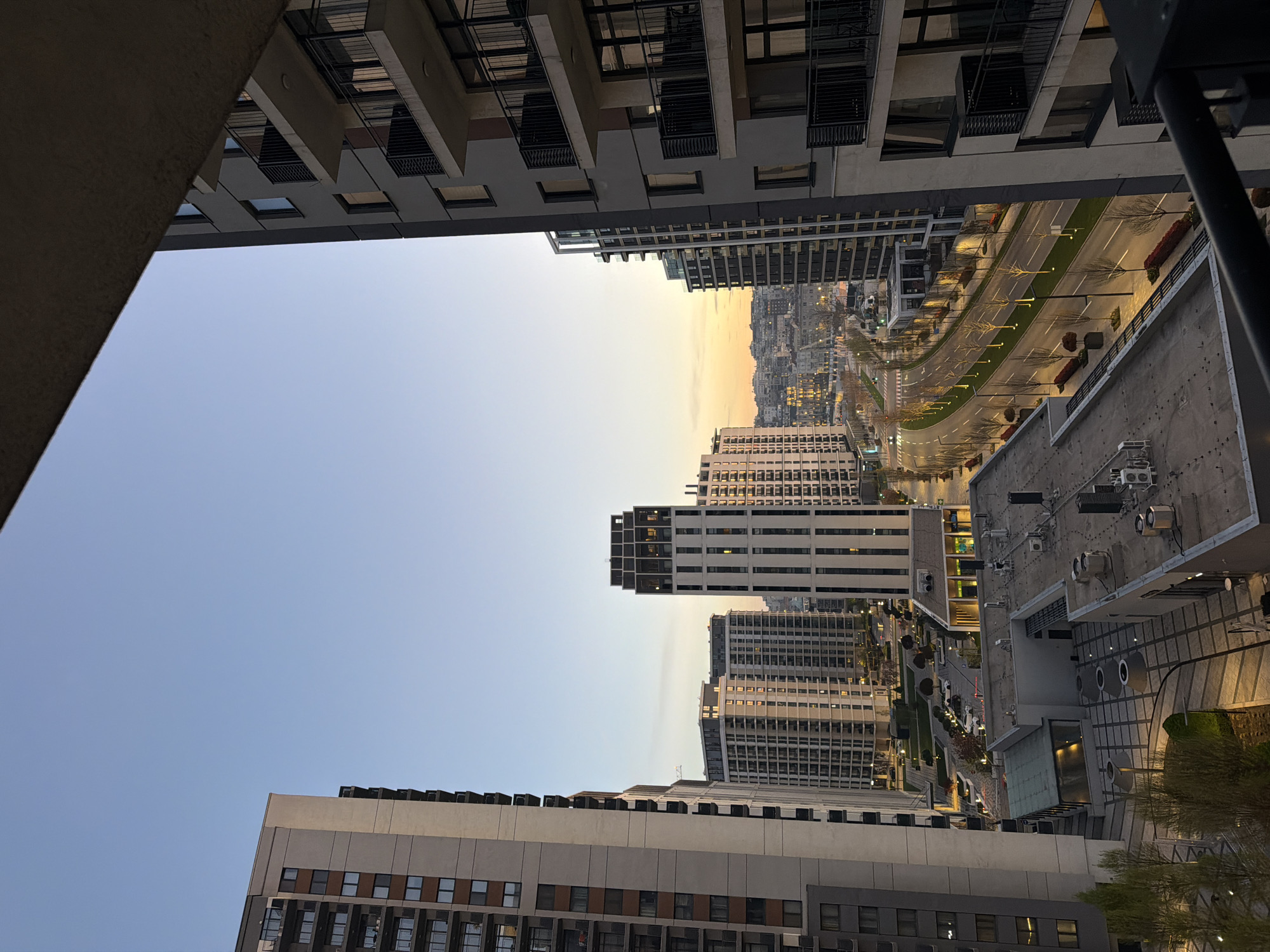 The image shows a cityscape in Belgrade, Serbia, viewed from a high vantage point, likely a balcony or window within a tall building. The perspective is slightly tilted, showcasing a mix of modern high-rise buildings and urban infrastructure. In the distance, a broad avenue is visible, lined with trees and streetlights. The lighting suggests either dawn or dusk, with a soft, gradient sky visible between the buildings. The architectural style of the buildings is predominantly modern, with clean lines and geometric shapes. No people are directly visible in the image. The environment is a densely populated urban area.