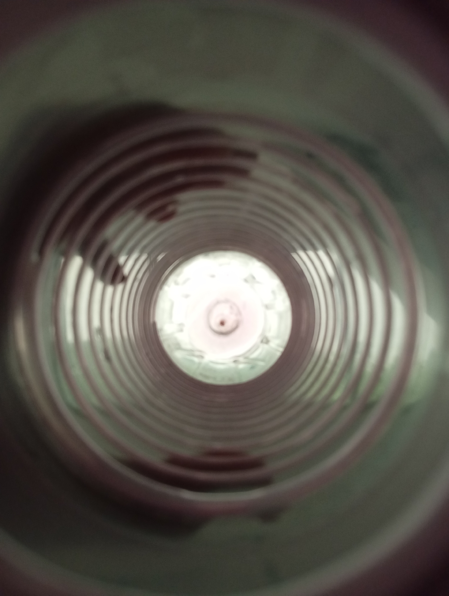 An extreme close-up shot looks down the inside of a ribbed, cylindrical object, possibly a pipe or bottle. The perspective is from within the object looking outwards, creating a tunnel effect. The ribbed interior is a dull reddish-brown color, with each rib casting a shadow. Towards the center of the frame, the opening of the cylinder is visible, revealing a bright, hazy light. The outer edges of the image are blurred and darker, suggesting the perspective is from deep within the cylinder, or the object itself is dark. There are no visible people or specific identifiable features within the scene that would allow for a location to be determined.