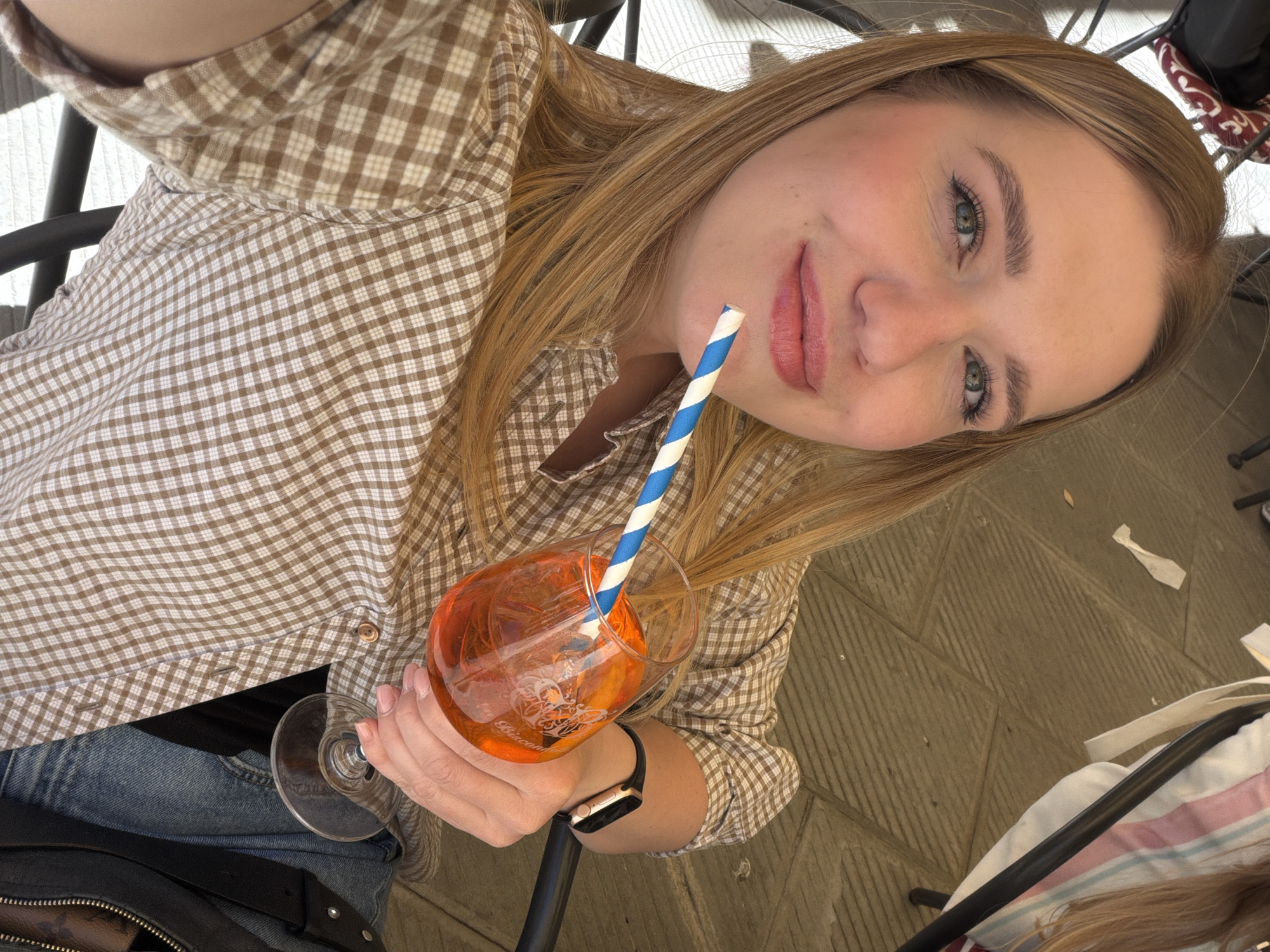 A young woman, possibly in her early to mid-20s, is taking a selfie while seated at a table. She is wearing a brown and white checkered shirt, jeans, and a black watch. She is holding a wine glass filled with an orange liquid and a blue and white striped straw. The background appears to be an outdoor seating area with stone flooring. The location may be San Gimignano, Italy, given the overall aesthetic.