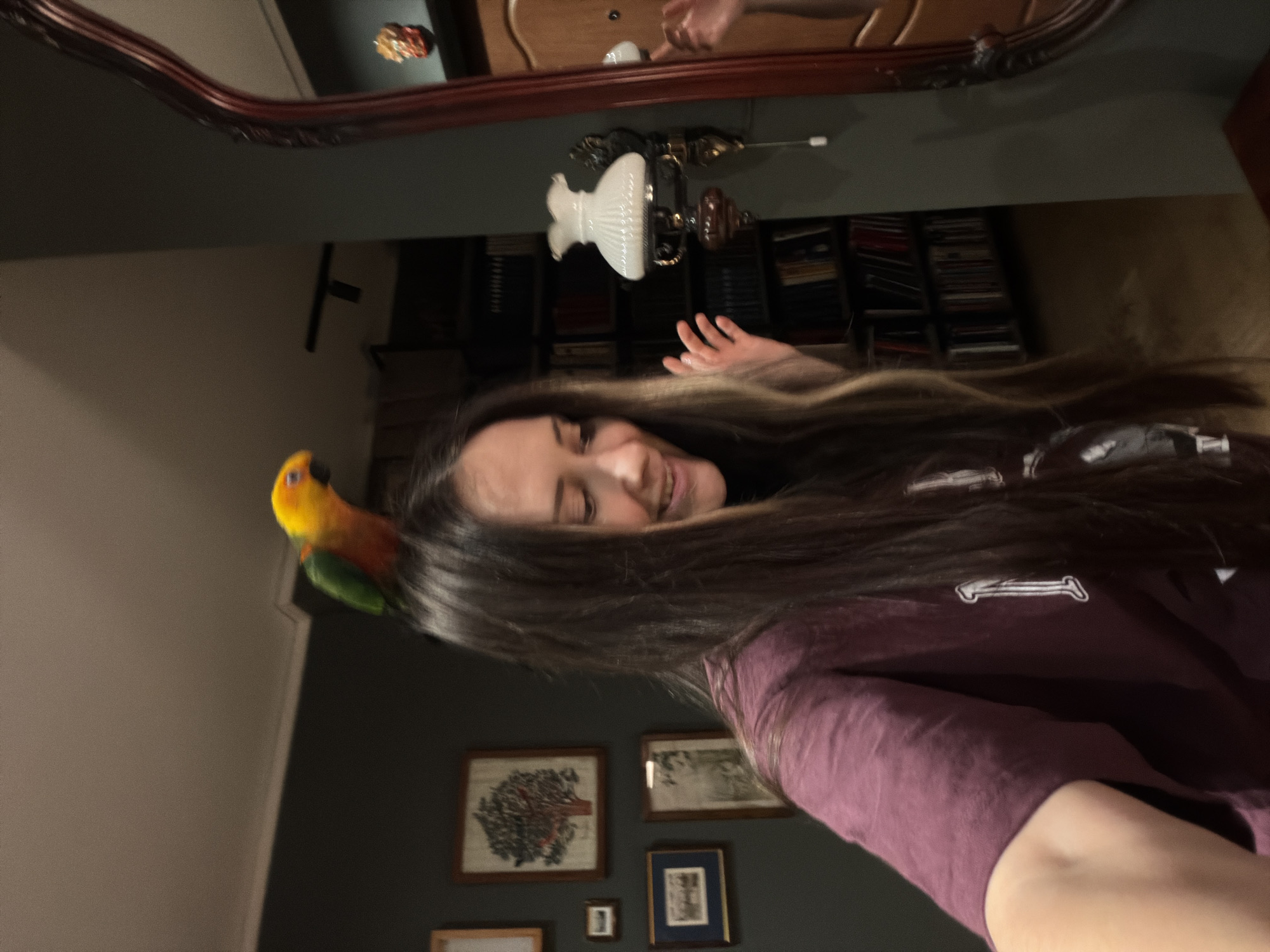 A woman with long brown hair has a sun conure parrot perched on her head. She is smiling and reaching upward with her right hand. The environment appears to be a room with a bookshelf, a lamp, a mirror, and framed pictures on the wall. There is no indication of the location of Khimki, Russia.