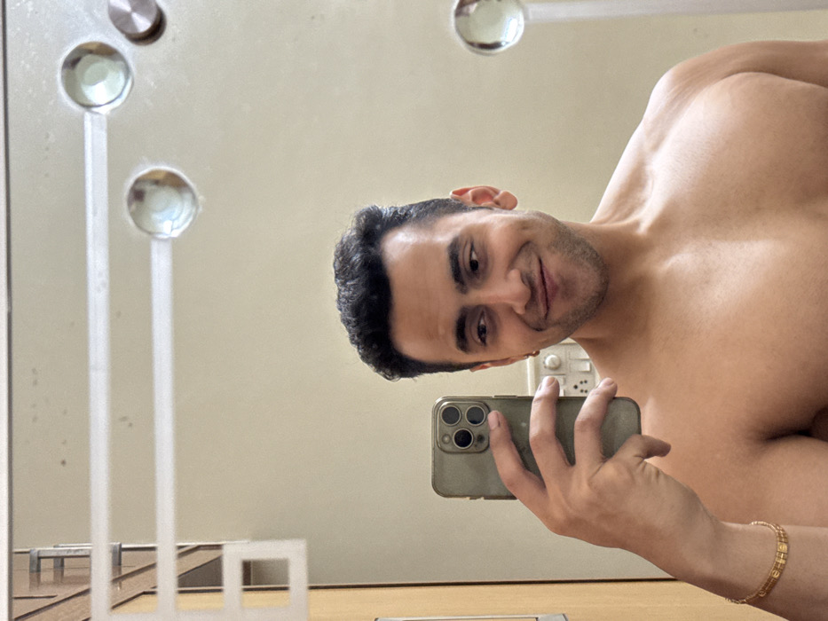 A mirror selfie features a shirtless male from the chest up, with dark, curly hair and stubble, looking at the camera with a slight smirk. He holds a light-colored smartphone, likely an iPhone due to its triple-lens camera module, in his right hand. A gold-colored bracelet with rectangular links adorns his left wrist.
The indoor setting, reflected in the mirror, includes a plain, light-colored wall. On the left side, two circular, clear, possibly glass or crystal, decorative elements are visible, each with a slender white vertical support. Another similar circular object is reflected higher in the upper left corner. Behind the man's right shoulder, a white electrical outlet or light switch panel is visible on the wall. Towards the bottom left, some geometric white structures, resembling a railing or decorative screen, are reflected on a wooden or beige-toned floor/countertop.
The lighting appears bright and diffused, suggesting daytime. The man is engaging directly with the camera for the selfie. There is no visible text in the image. The provided location context is Kolkata, India.