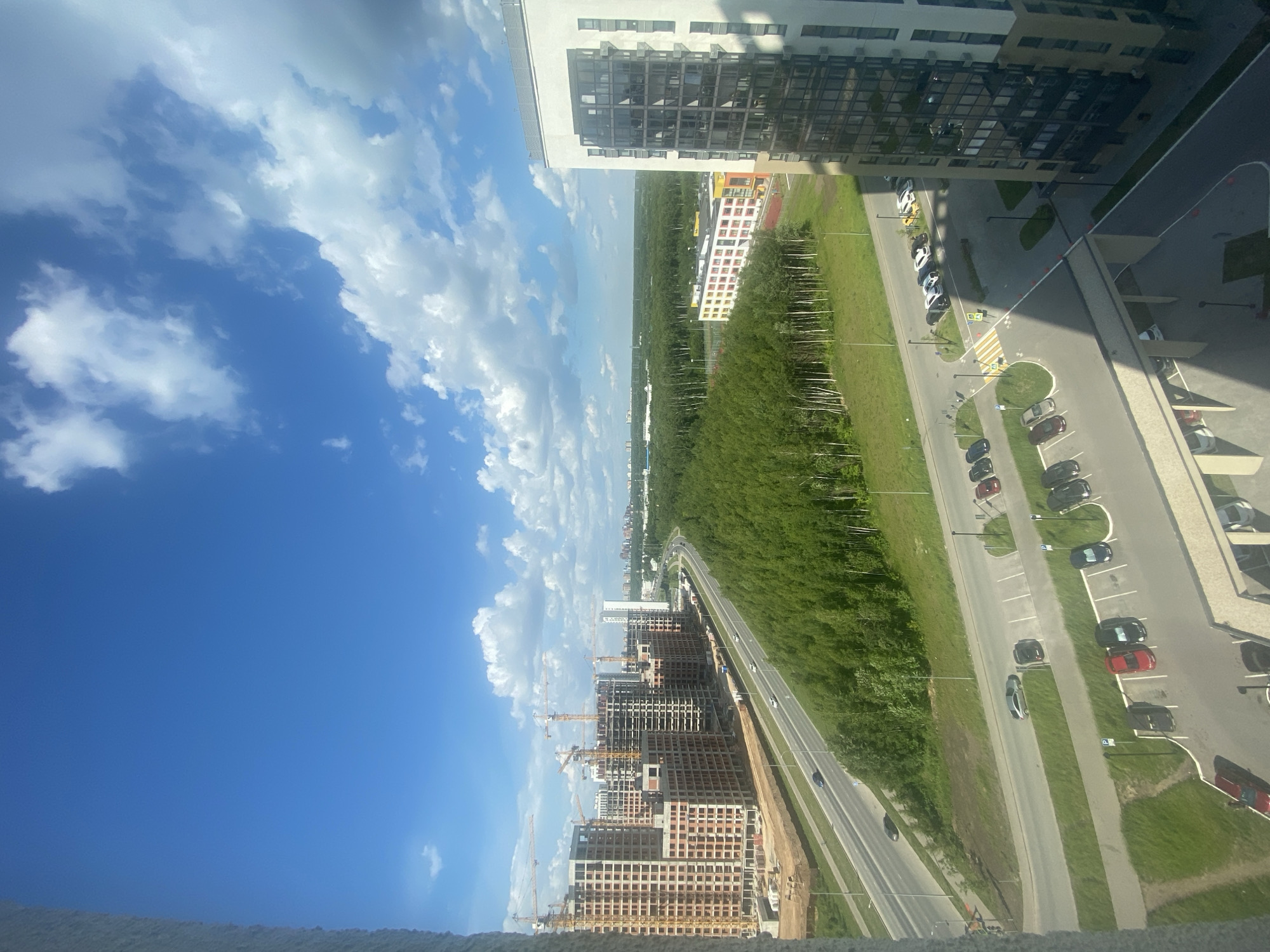 The image captures a wide aerial view of a developing urban area on a bright, sunny day in Kazan, Russia. The foreground is dominated by a large, multi-story residential building under construction on the left, with several construction cranes visible. Adjacent to the construction site is a multi-lane road with a few cars driving. Beyond the road, a dense green forest stretches across the landscape. In the middle ground, there is a newly constructed school building with a colorful facade, and further in the distance, more residential buildings and infrastructure can be seen, hinting at further development. The sky is a vibrant blue with scattered white clouds. The scene suggests a modern city expanding into a green, natural area.