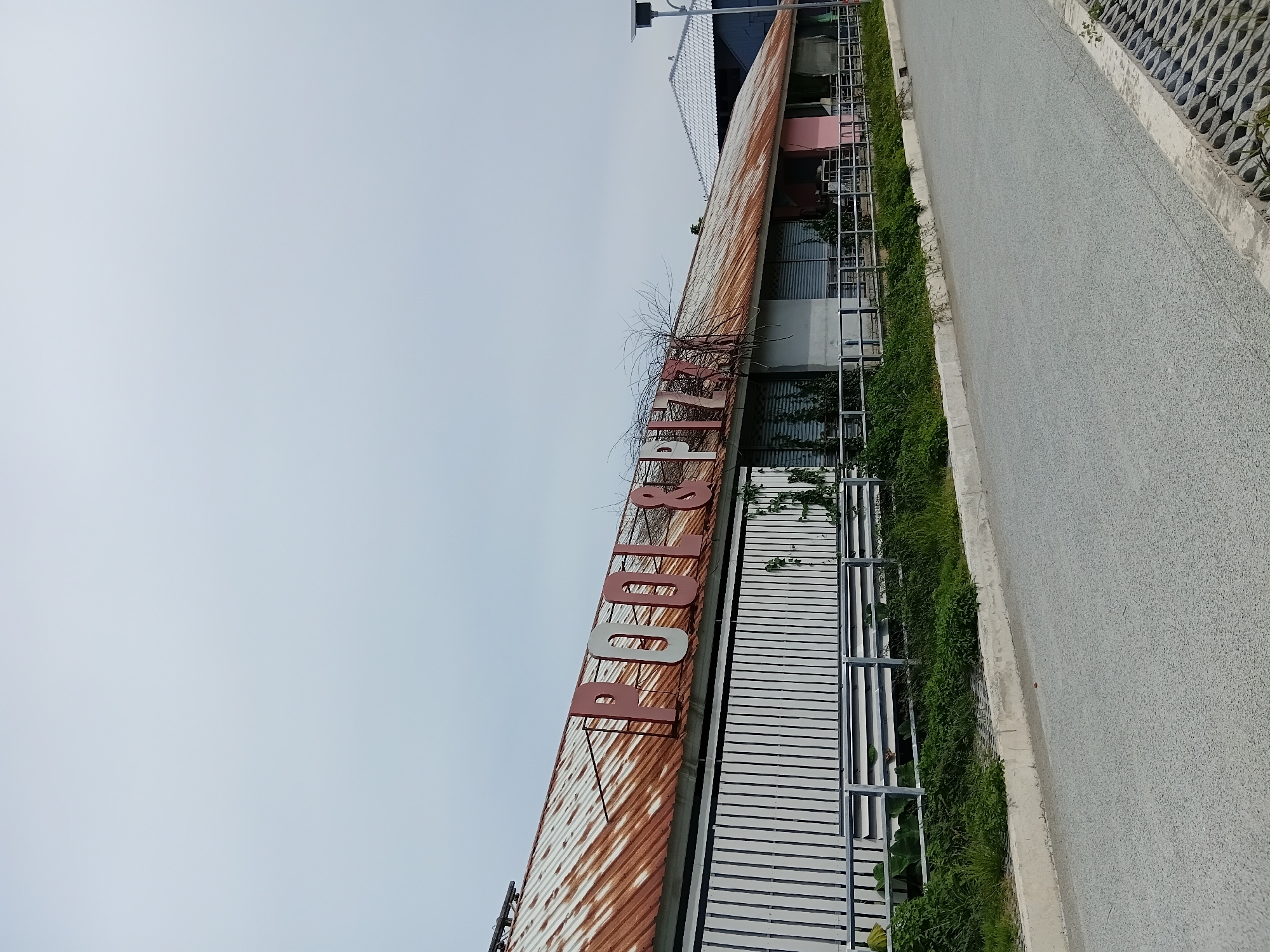 The image depicts a run-down building with a rusted corrugated metal roof. Large, red and white letters spelling "POOL & PIZZA" are affixed to the roof. The building has a white slat-like facade and several closed shutters. Greenery, including vines, grows around the lower part of the building and along a metal fence. A paved road is visible in the foreground, with a concrete curb and some decorative concrete blocks on the right side. The sky is overcast and pale. The scene appears to be in a semi-urban or commercial area, possibly in Ko Samui District, Thailand, given the architecture and vegetation. No people are visible in the image.