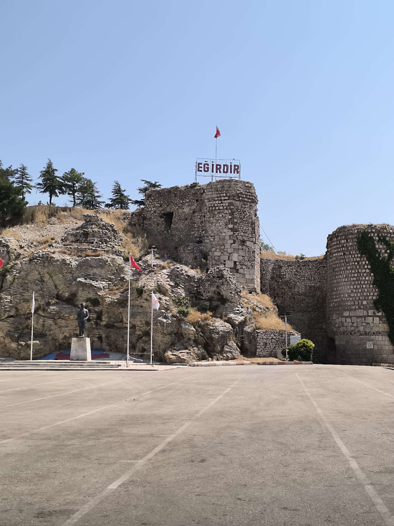 A statue of a man stands on a pedestal in front of a rocky hill. The hill has ruins of a castle on top, with a sign that says "EĞİRDİR." The castle is made of stone and some parts are covered in ivy. In front of the hill, there is a paved area with parking lot lines. Several flags are flying on poles. The sky is clear and blue. The environment appears to be an outdoor public space in the city of Eğirdir, Turkey.