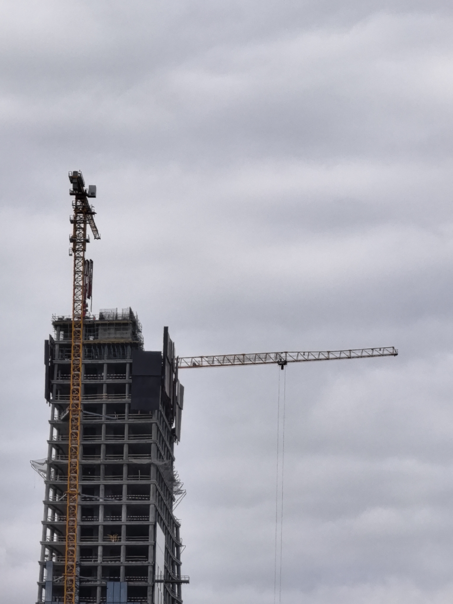 An overcast sky serves as the backdrop for a high-rise building under construction in the city of Unknown, Unknown. A large yellow construction crane is attached to the side of the partially built skyscraper. The building itself is a skeletal structure of concrete and rebar, with multiple floors visible. Another, longer crane extends horizontally from the side of the building, reaching out into the sky. The image captures a moment in the ongoing process of urban development.