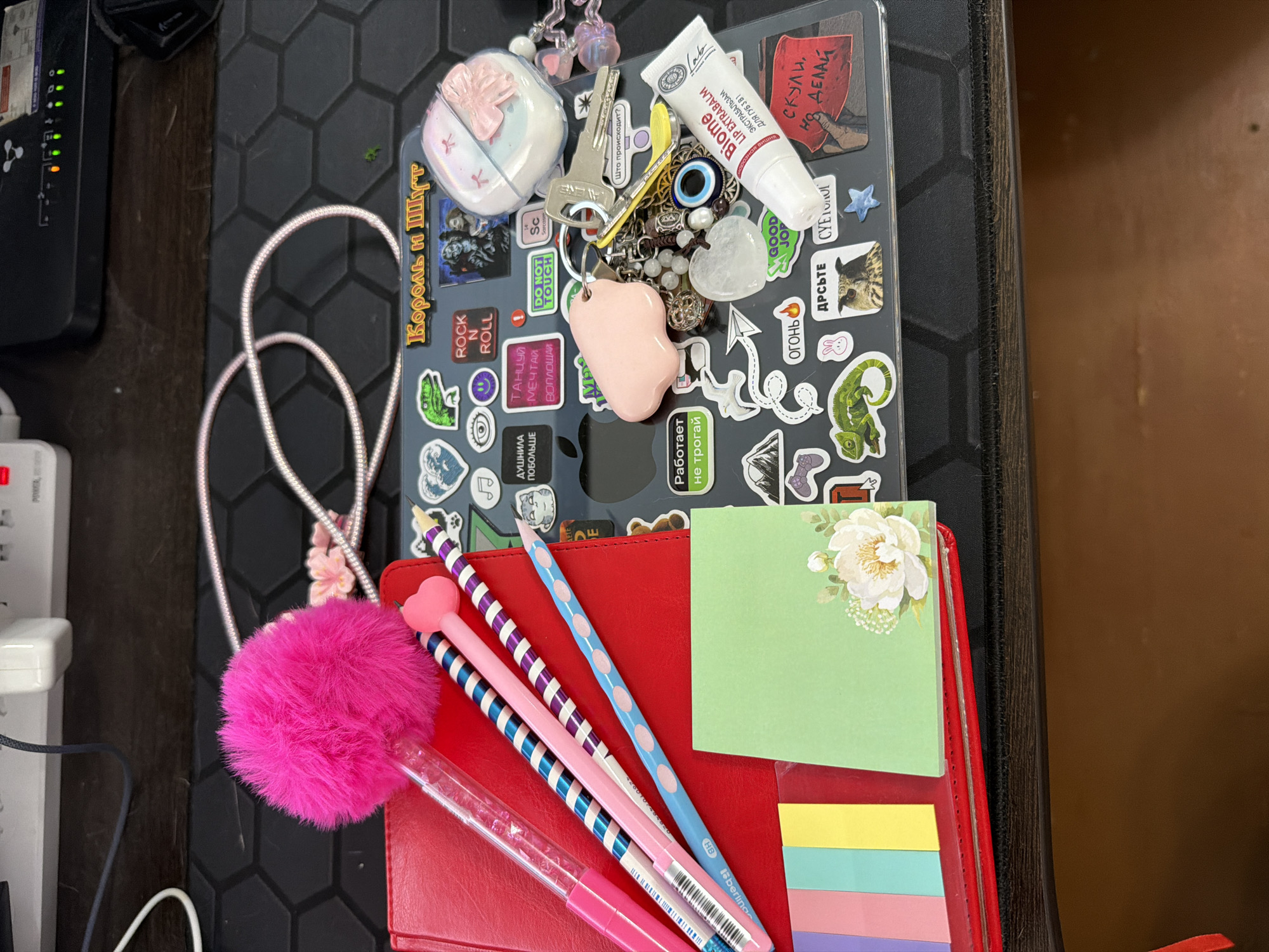 The image displays a cluttered desk surface, possibly in Chaikovsky, Russia, with a variety of personal items. A red notebook is visible on the left, topped with a bright pink fluffy pom-pom pen and several other pens with patterned barrels in pink, blue, and white. To the right of the notebook, a green sticky note with a floral illustration is placed. A significant portion of the image is occupied by a laptop or tablet, adorned with numerous stickers. These stickers include text in Cyrillic, a green lizard, an owl, a dinosaur, hearts, and various other graphics. Among the items on top of the laptop are a set of keys, a pink charm shaped like a droplet, several beaded bracelets, and a tube of lip balm labeled "Biome LIP EXTRA BALM." The background shows a dark, textured surface, possibly a desk mat, with a white power strip and electronic devices mounted on a wall in the upper section. The overall impression is one of a personal workspace with decorative and functional items.