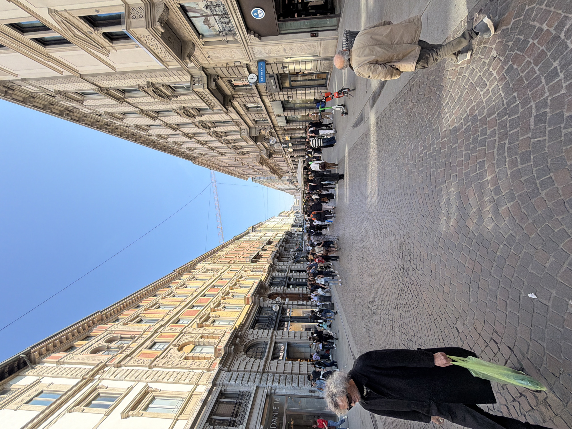 The image is a ground-level view of a street in Milan, Italy, with tall buildings lining each side, converging in the distance. The street is paved with cobblestones. A queue of people stretches along one side of the street. Individuals are walking on both sides, including an elderly person with gray hair in the foreground holding a green bag and another wearing a tan coat further along the street. The sky is clear and blue. A construction crane is visible in the distance. Signage on the buildings includes the names "NORTH SAILS" and "PAUL SHARK" along with 'DANTE 16'.