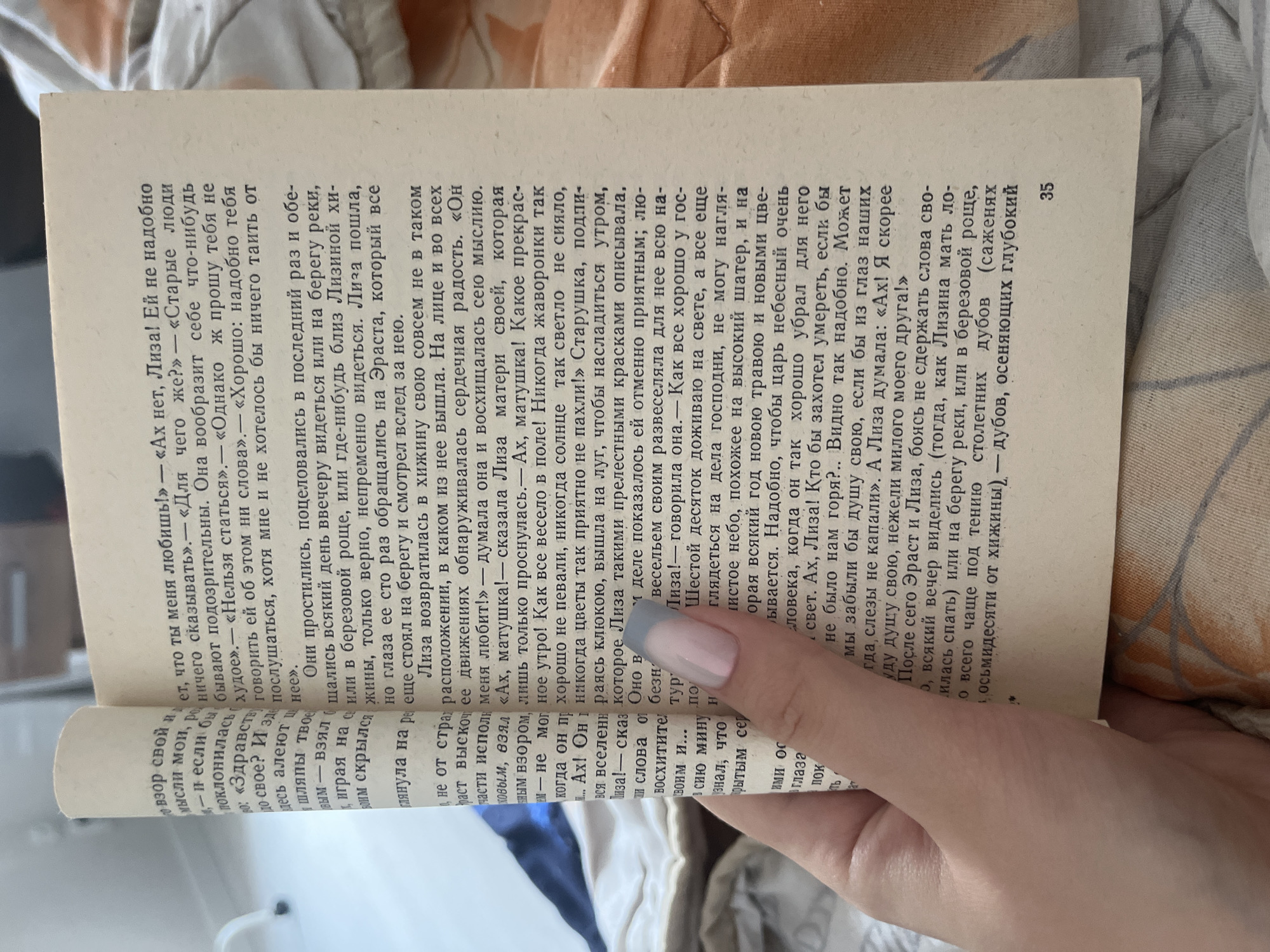 The image shows a close-up of an open book with Russian text. The book is a paperback with aged, yellowed pages. The text is printed in a serif font. The page on the right has a page number "35" at the bottom right. A hand with manicured nails is holding the book open, with the thumb and index finger near the spine on the left page. The background is out of focus and appears to be a textured surface, possibly fabric. The lighting suggests an indoor setting. There is no information in the image to indicate a location in the city of Perm, Russia.