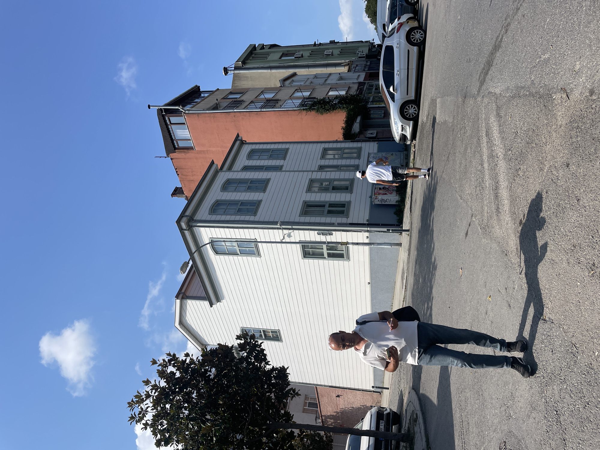 A man wearing a white polo shirt and blue jeans walks on a street in Kadıköy, Turkey, holding a phone and a small card. He is carrying a dark bag over his right shoulder. Behind him, another person in a white t-shirt and shorts walks away from the camera, wearing a white cap. The street is paved and appears to be a residential or mixed-use area.
Several buildings line the street. To the left of the foreground man is a white, two-story house with light blue trim around its windows. Adjacent to it are taller, multi-story buildings with orange, gray, and light green facades. The orange building has several balconies and a dark awning over a window on the top floor. The gray and green buildings also feature balconies and appear to be older structures.
In the background, a large tree with dark green leaves stands to the left of the white house. The sky is a clear blue with a few scattered white clouds. Several cars are parked or driving on the street, including a white Peugeot hatchback in the middle ground and a white SUV on the left. The overall scene depicts a sunny day in an urban environment.