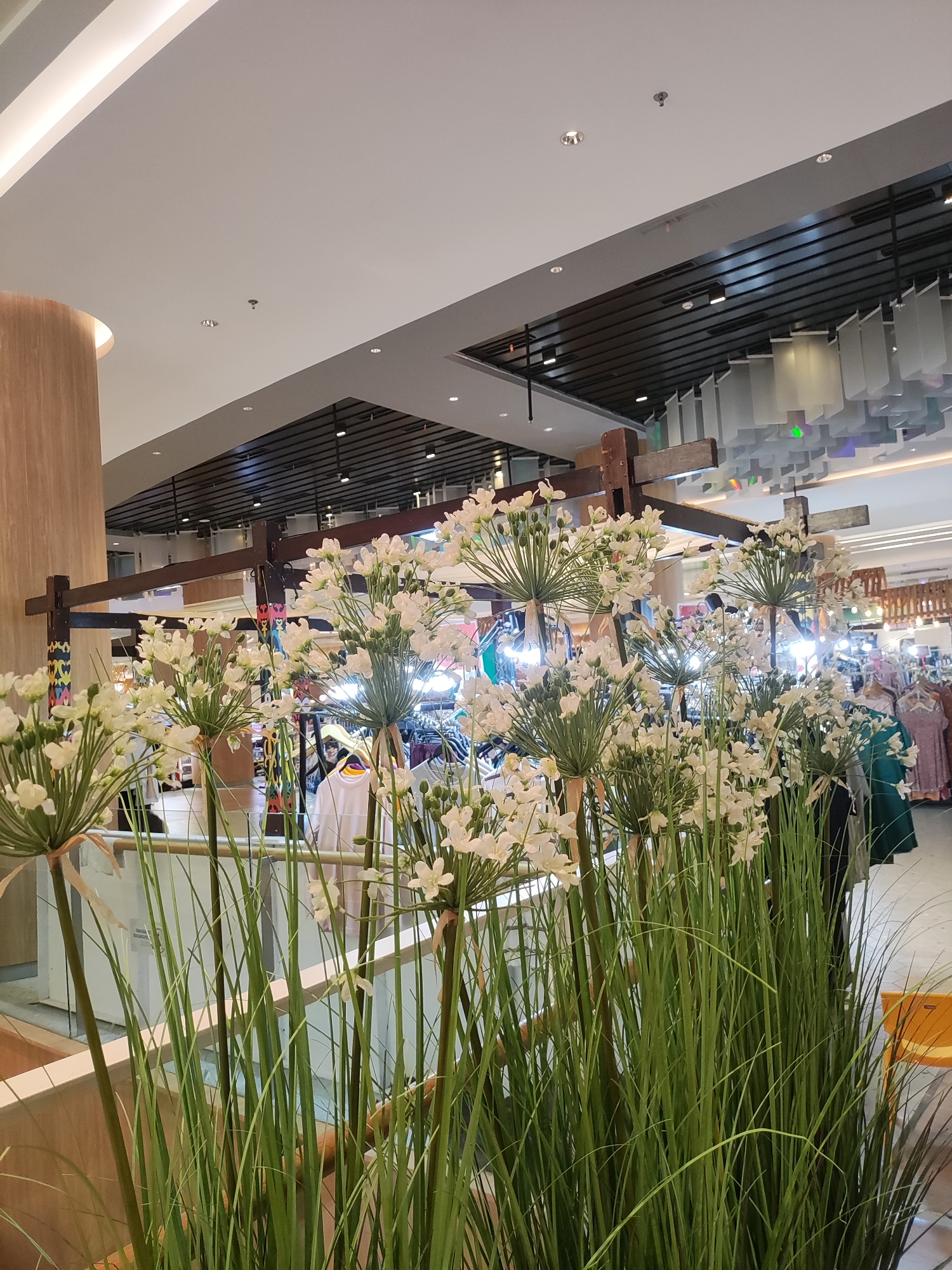 The image depicts an indoor scene, possibly a shopping mall or a department store, with a prominent display of artificial white flowers and tall green grass in the foreground. These decorative elements are arranged in front of a retail environment. In the background, racks of clothing are visible, suggesting a clothing store. People are present in the background, browsing or working in the store. The ceiling features modern lighting fixtures and architectural elements. The overall ambiance is that of a commercial space. The image does not provide specific details about the city of Lambangjaya, Indonesia, or any unique landmarks related to that location.
