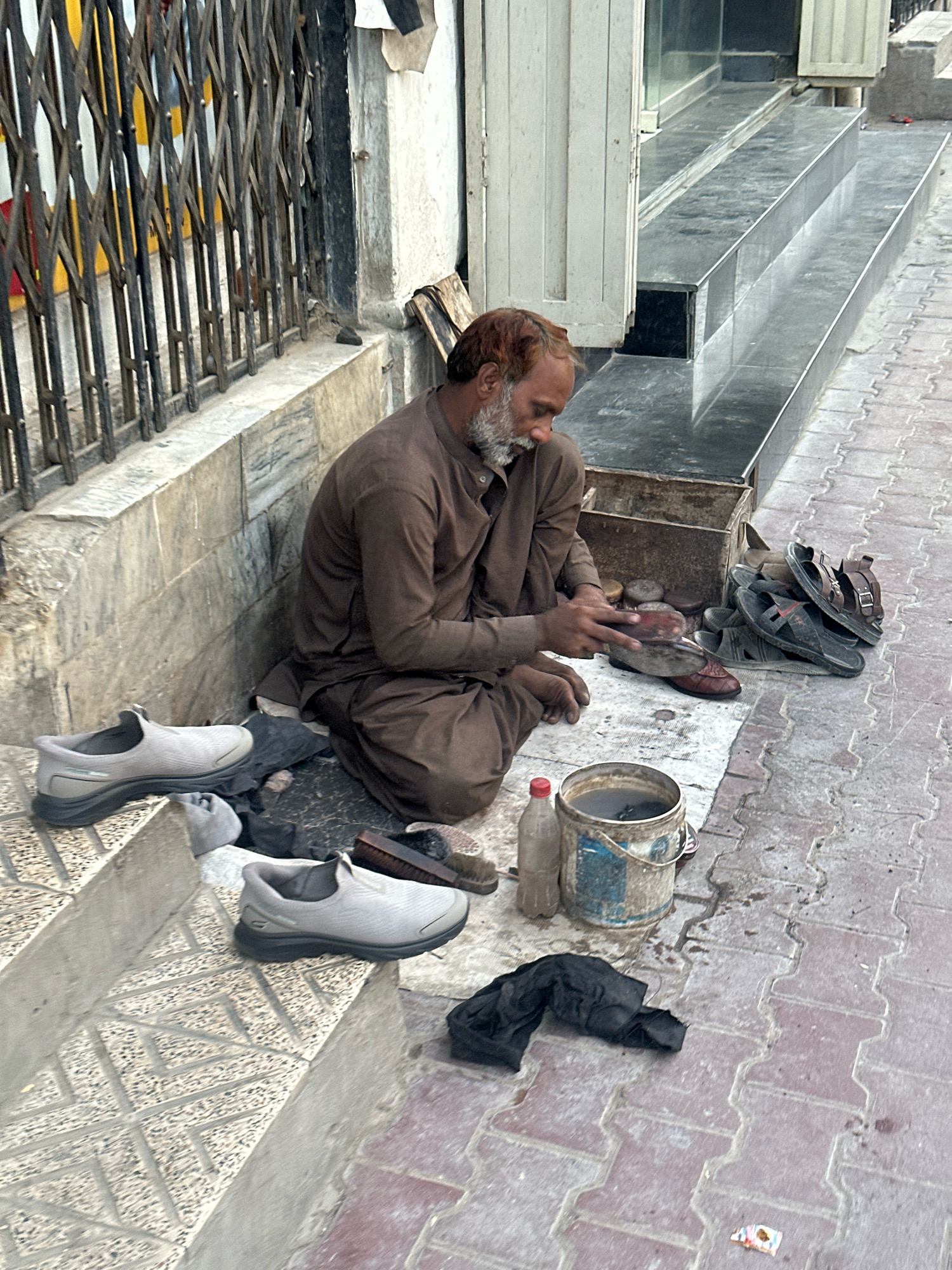 A man is sitting on the ground in front of a building, performing shoe repairs on a sidewalk in Quetta City Tehsil, Pakistan. He is wearing a brown garment and has a grey beard. To his left are several pairs of shoes, including two grey sneakers on a stone step. In front of him is a metal bucket filled with water, a plastic bottle, and various shoe repair tools and materials. A pile of dark fabric is on the ground to his right. Behind him is a metal grate gate and a stone wall. The sidewalk is paved with reddish-brown interlocking bricks.