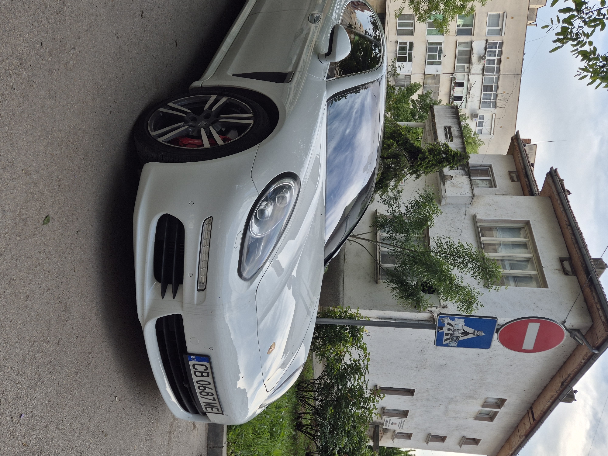 A white Porsche Panamera is parked on the street in Pavlikeni, Bulgaria. The car has a Bulgarian license plate with the characters "CB 0687 ME". A pedestrian crossing sign and a "no entry" sign are posted on a pole adjacent to an old building. A multi-story residential building is in the background. Vegetation is visible around the street.