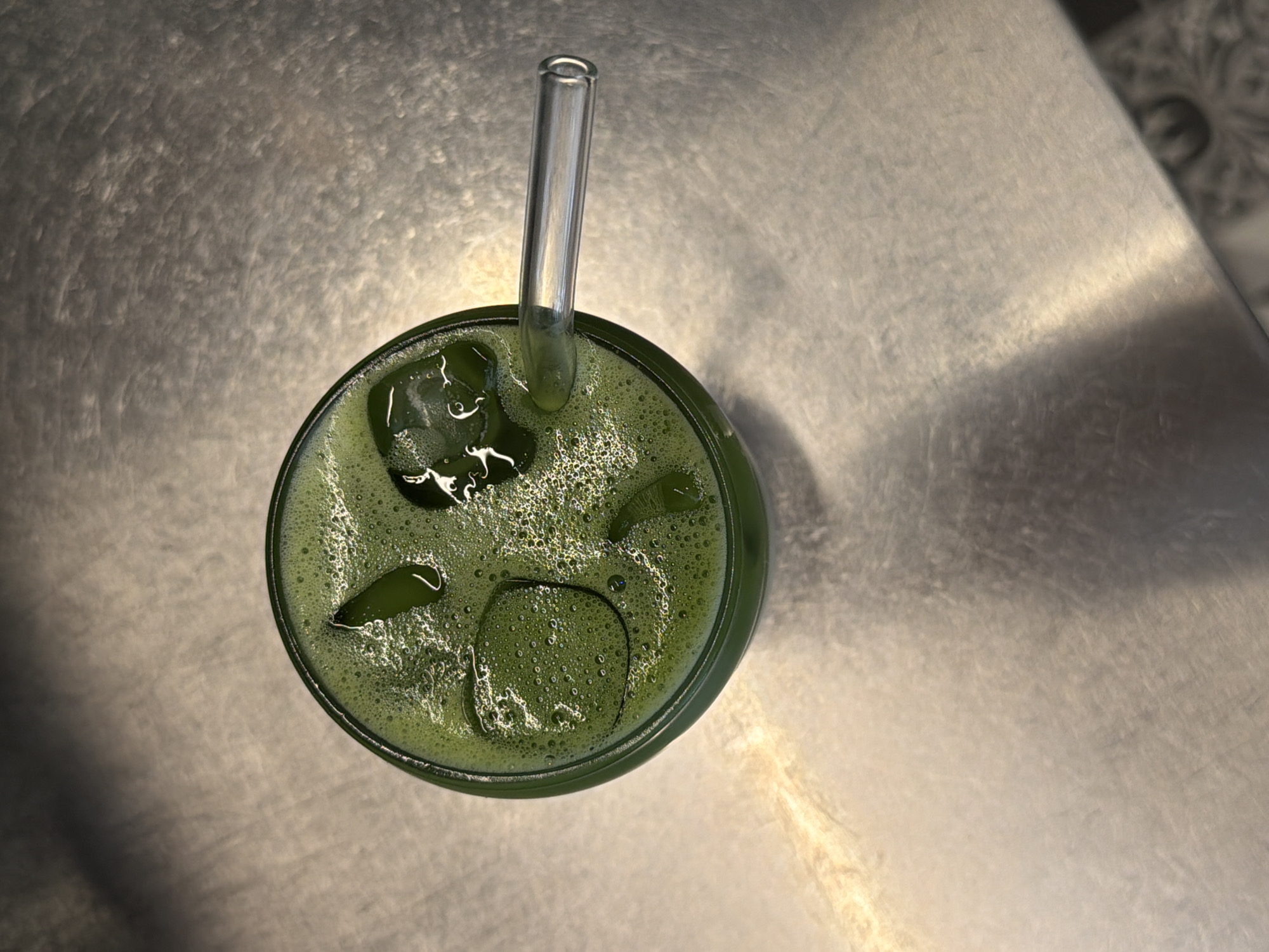 A glass of green juice with ice and a straw rests on a metallic surface. The glass is round with a green rim. The juice is a vibrant green and frothy, with several ice cubes visible. A clear glass straw extends from the right side of the glass into the liquid. The surface beneath the glass appears to be brushed metal, with visible scratches and reflections. Shadows are cast across the surface, suggesting overhead lighting. The image is a top-down view. The location in the city of Sofia, Bulgaria, is not discernible from the provided image.
