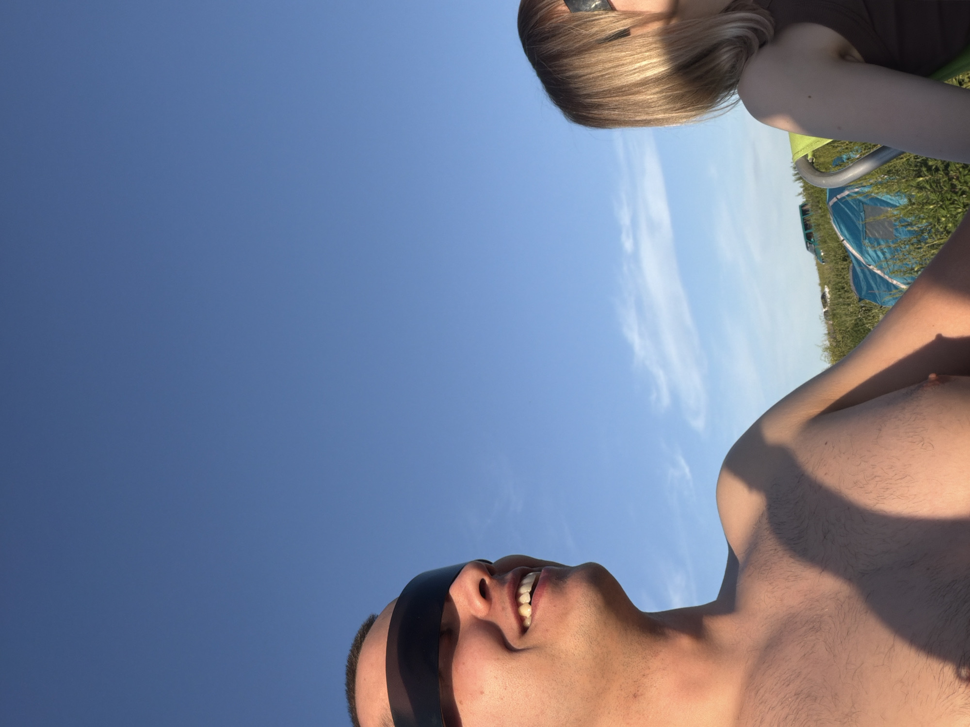 The image shows two people in an outdoor setting, likely a field or park, under a clear blue sky with some wispy clouds. The person on the left is a male, shirtless and wearing large black sunglasses that cover his eyes. He is smiling and looking to the right. His chest and shoulder are visible, with some light chest hair. The person on the right is a female, with blonde hair and also wearing sunglasses. She is partially visible from the shoulder up, with her head turned to the left. In the background, there are some tents and possibly a vehicle, suggesting a camping or outdoor event. The overall lighting suggests it is daytime, possibly late afternoon due to the warm tones on the skin. The location is identified as Povarovka, Russia.