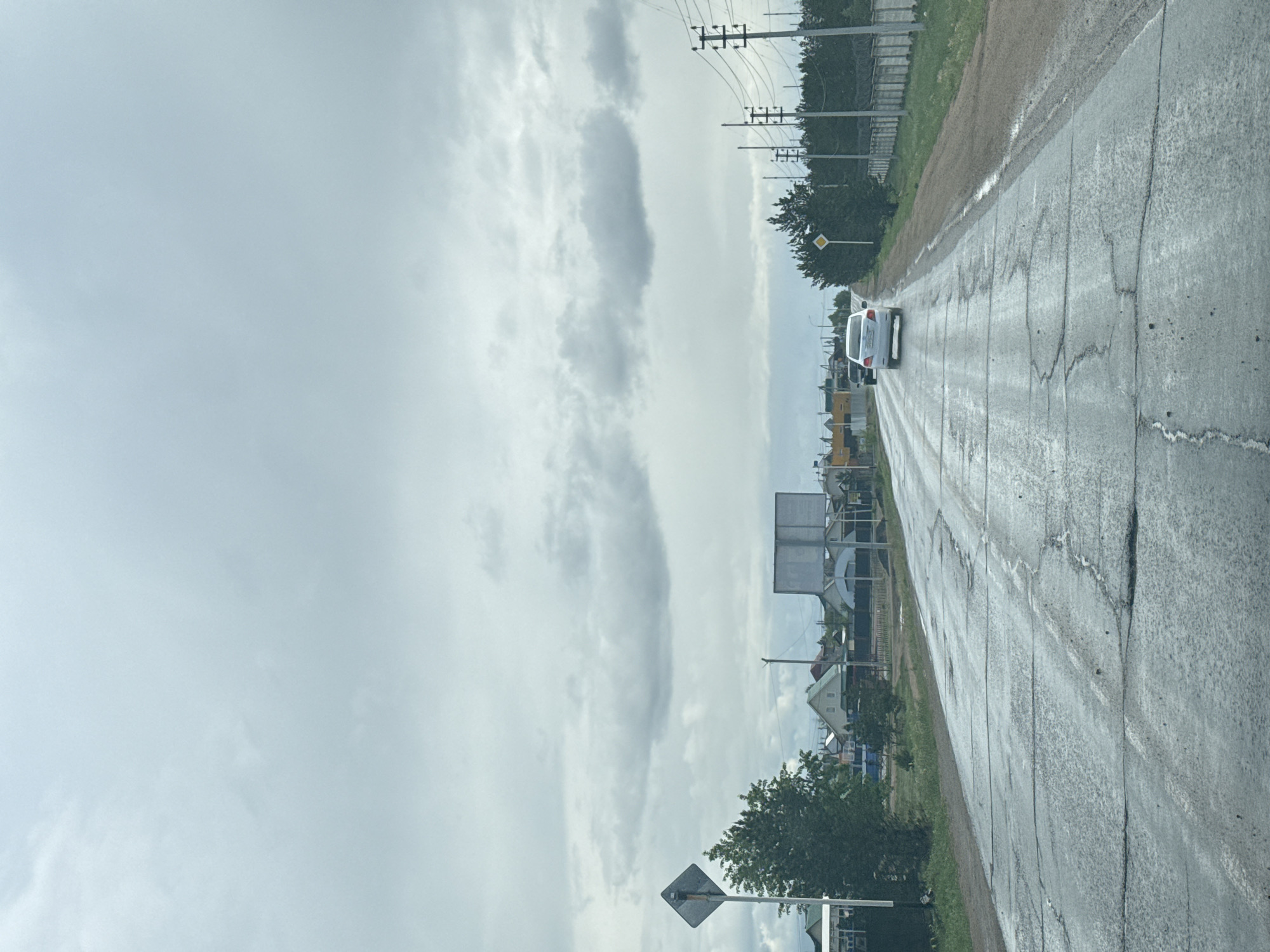 The image depicts a straight asphalt road viewed from inside a vehicle, possibly taken from the front passenger seat or dashboard. The road is wet, indicating recent rain, and shows visible cracks and patching. A light-colored SUV or minivan is driving ahead in the same direction.
The sky is overcast with various shades of grey clouds, suggesting a cloudy day, possibly just after a rainfall. On the left side of the road, power lines and utility poles run parallel to the road. Further in the distance on the left, several buildings, possibly houses or light industrial structures, are partially visible through trees and behind a large, blank rectangular billboard structure. A yellow diamond-shaped road sign with a black border is also visible on the left side of the road, positioned near some trees.
On the right side of the road, there is a narrow dirt or grass shoulder, followed by more trees and vegetation. A fence line is visible above the trees further down the road on the right. The overall setting appears to be a semi-rural or exurban area. There is no discernible text on any signs or objects.