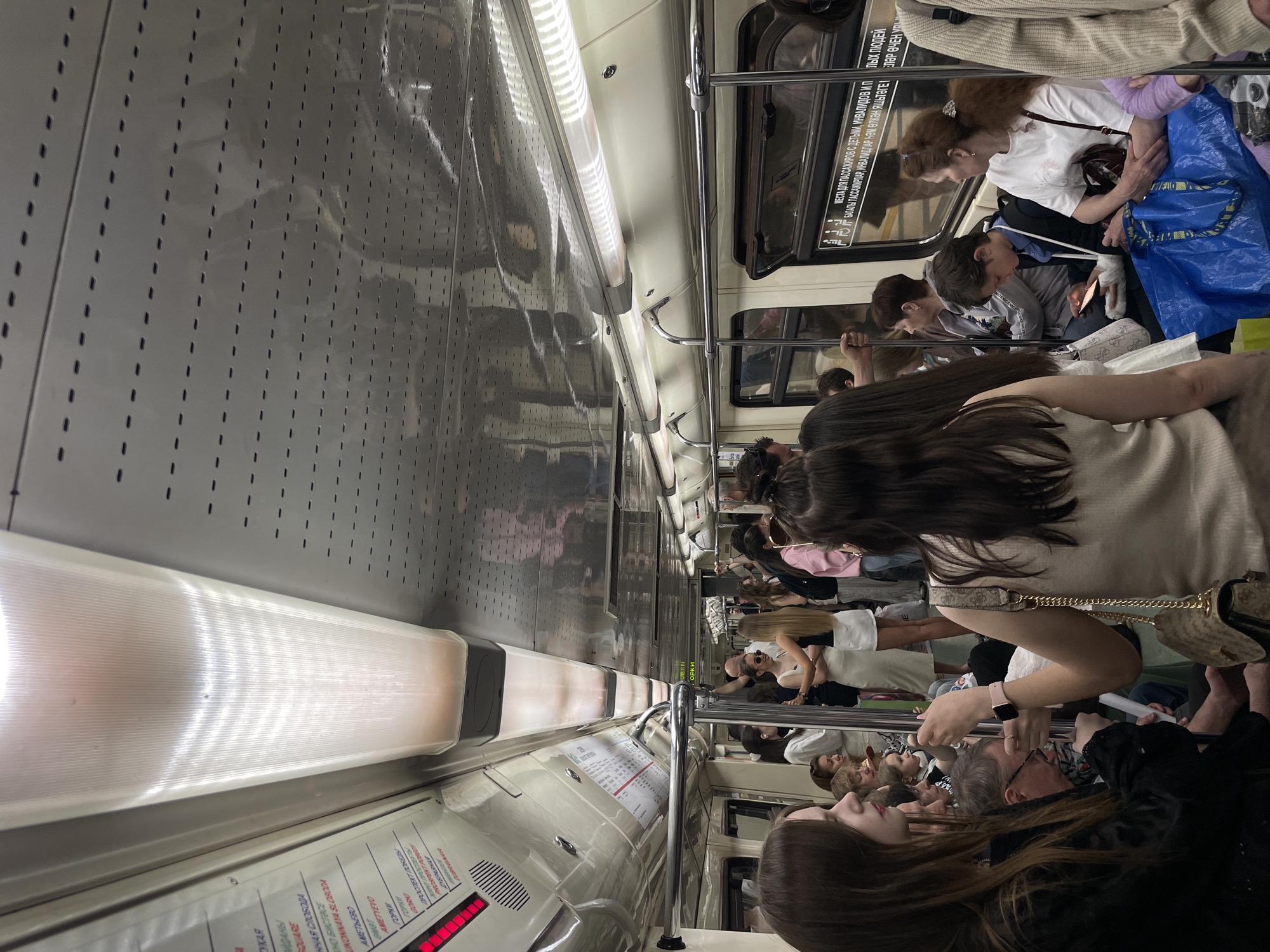 The image shows the interior of what appears to be a subway car, packed with people, likely during a busy commute. The interior walls of the car are visible, featuring rows of small perforations and informational signage in both Russian and another language, possibly Tatar. The signs mention stations like "Gorki", "Ametyevo", and "Prospekt Pobedy", indicating that this might be a metro system in a location where both languages are spoken. The car is filled with passengers of various ages and genders, some standing and holding onto the handrails. Details such as clothing, hairstyles, and the presence of personal belongings like bags provide a sense of realism and depict an everyday urban scene. The overall lighting is somewhat dim, typical of subway cars. The scene is crowded, suggesting a high level of transit use.