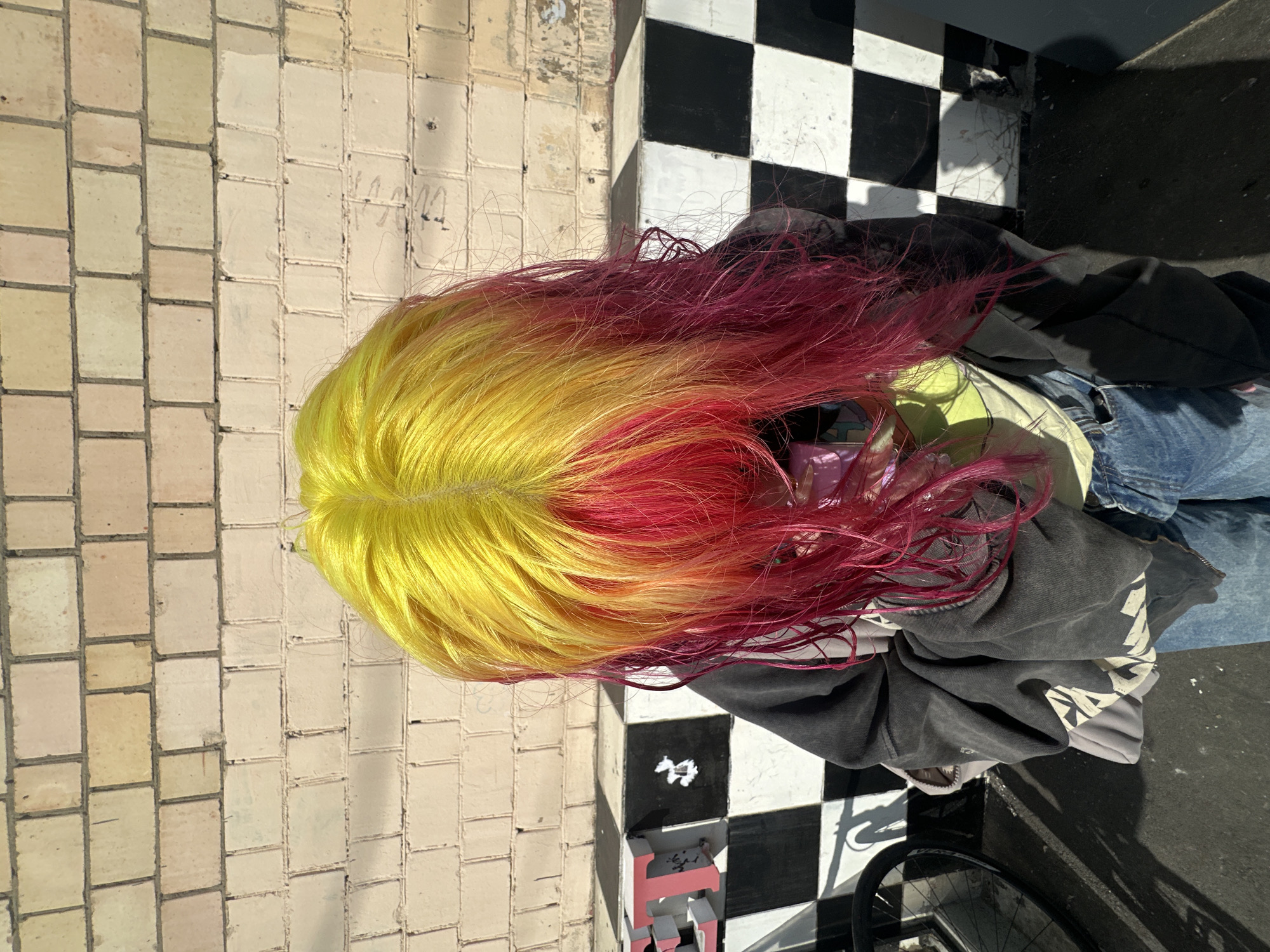 The image shows a person with bright yellow hair on top and pink hair underneath, standing outside. The background consists of a light-colored brick wall and a black and white checkered pattern on a lower surface. There are shadows on the ground, suggesting it is daytime. To the left, part of a bicycle wheel is visible. The person is wearing a dark hooded sweatshirt and blue jeans. The overall scene appears to be in an urban outdoor setting. There is no information in the image to identify the location as Moscow, Russia.